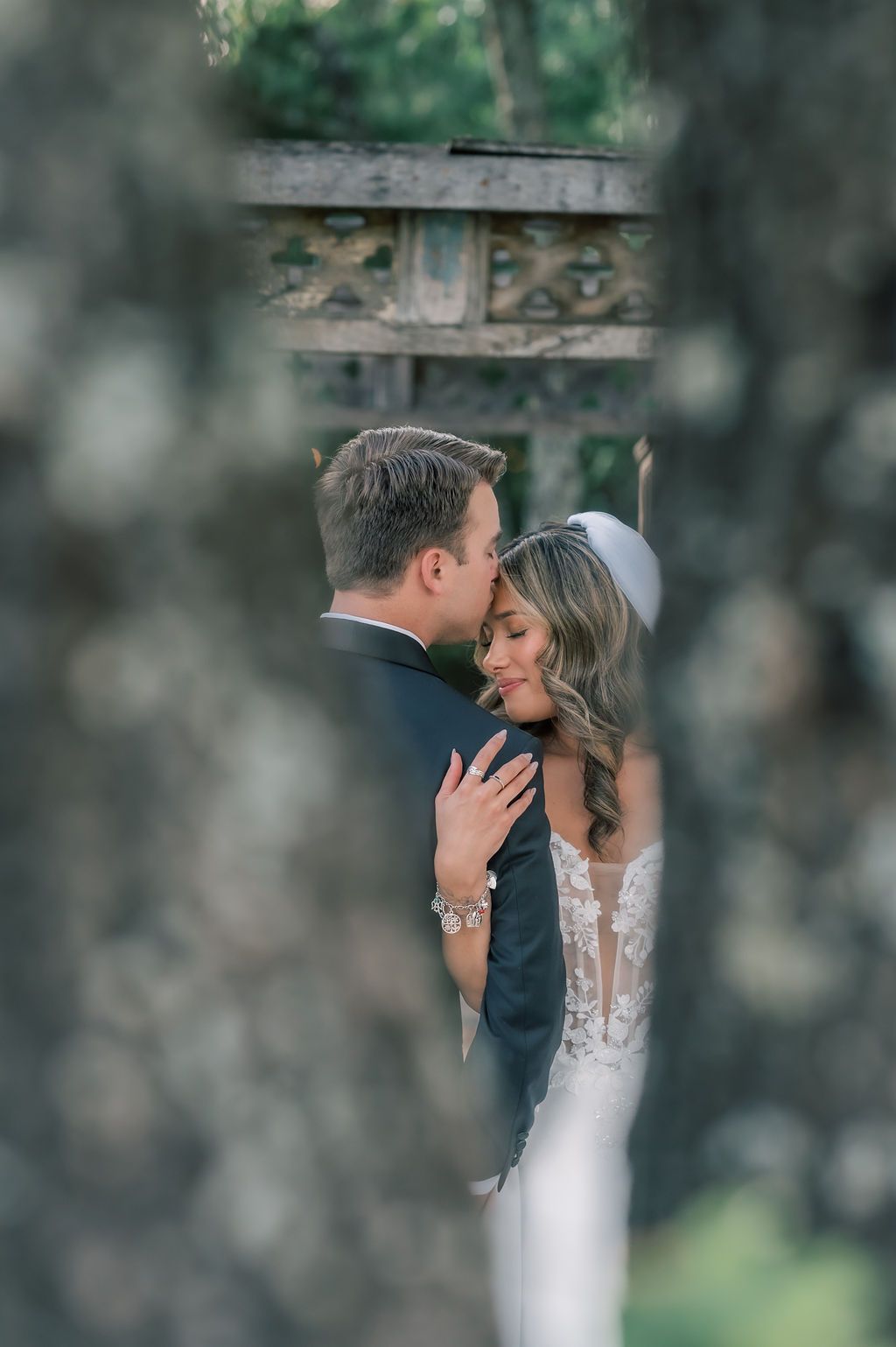 Bride and groom share a romantic embrace framed by moss-covered stone columns in an outdoor garden setting.