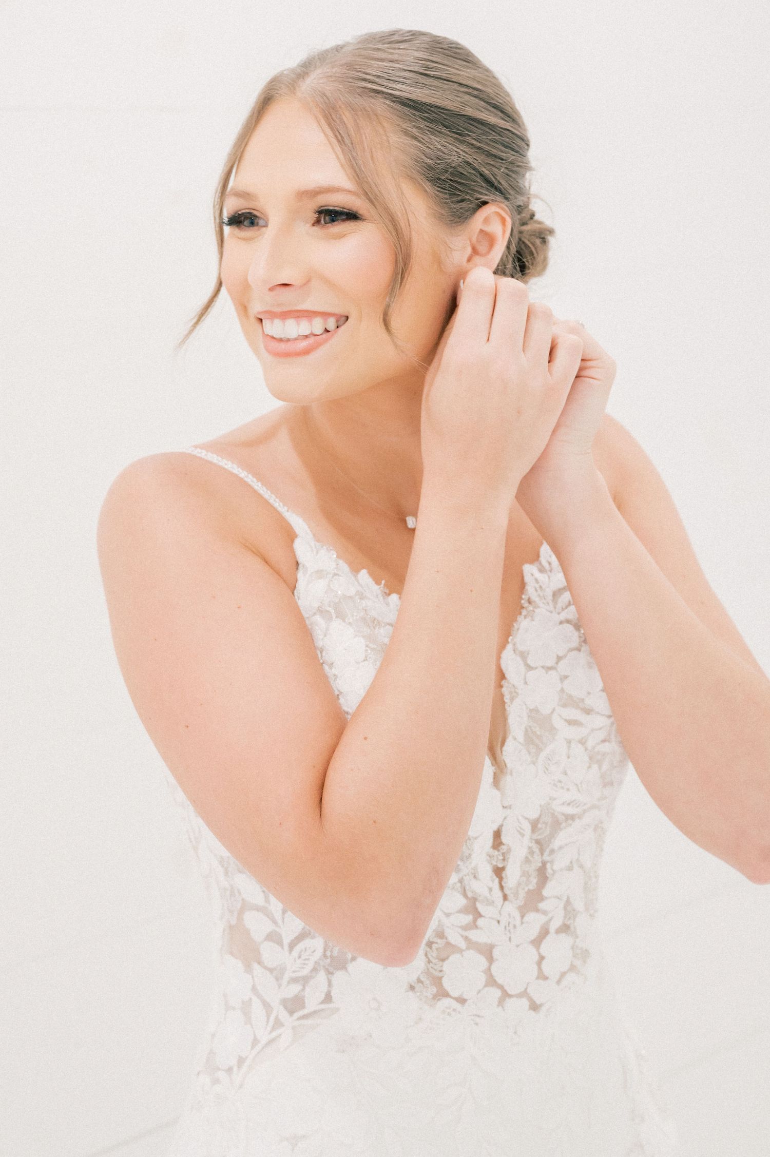 A bride wearing a delicate lace wedding dress smiles while putting on earrings against a white background.