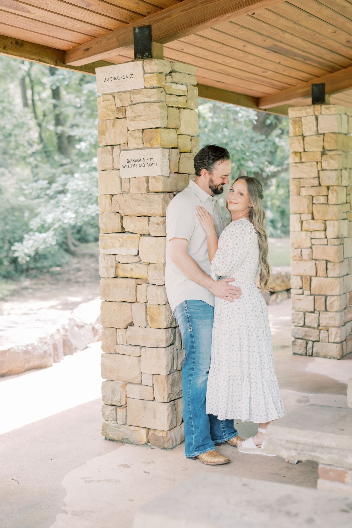 Couple embracing between stone columns under a wooden pavilion surrounded by lush green trees.