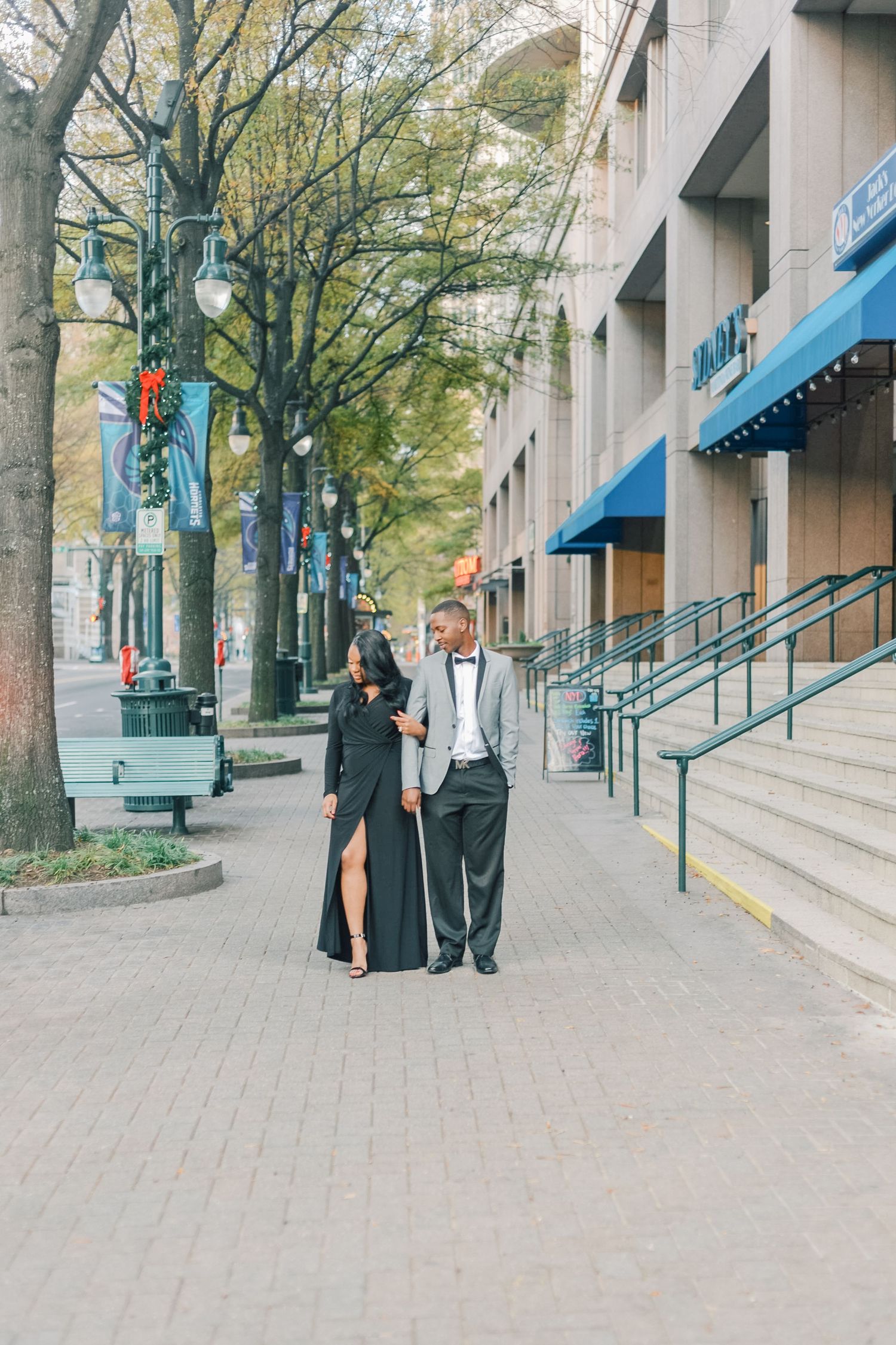 Couple in elegant formal wear walking together on a tree-lined city sidewalk with holiday decorations.