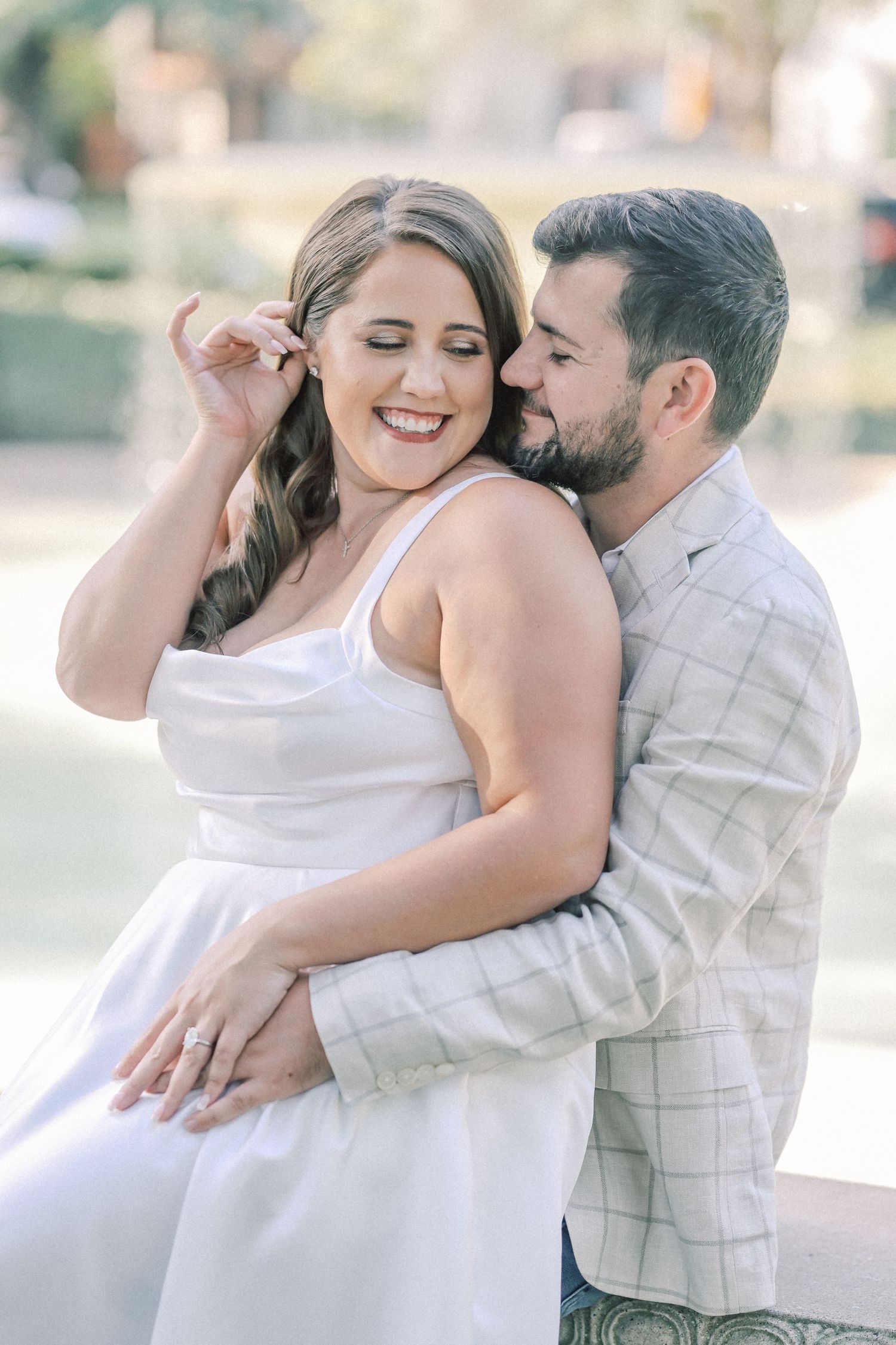 Laughing woman in a white satin dress tucks her hair behind her ear as her bearded fiancé embraces her from behind, ring on her hand.