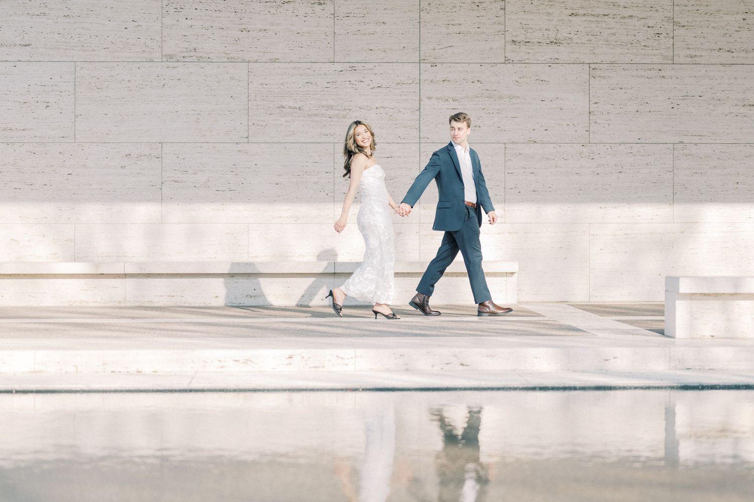 A couple in wedding attire walks together across a bright concrete plaza casting long shadows on the ground.