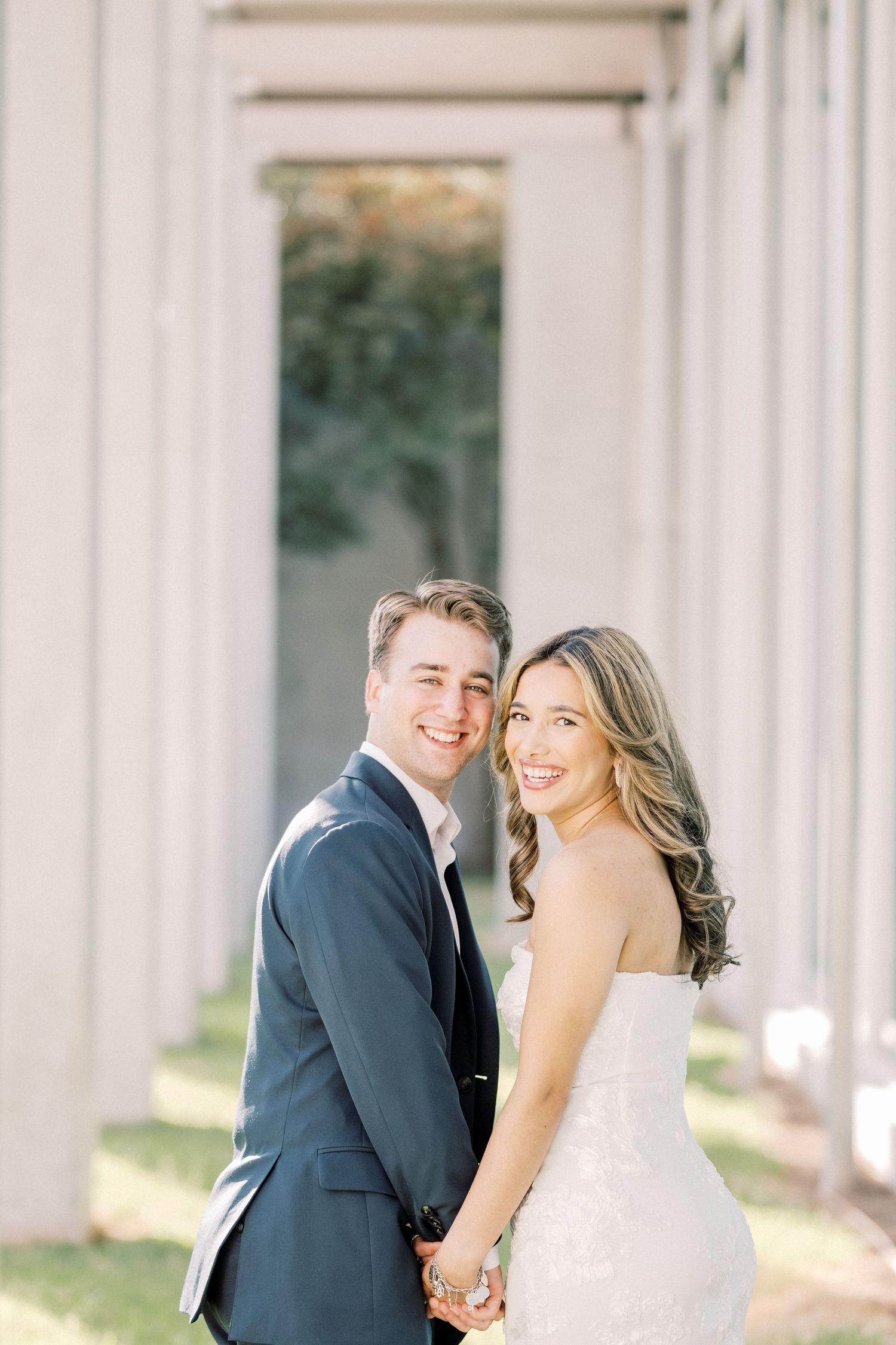 A couple shares a joyful moment together in their wedding attire among white columns on a sunny day.