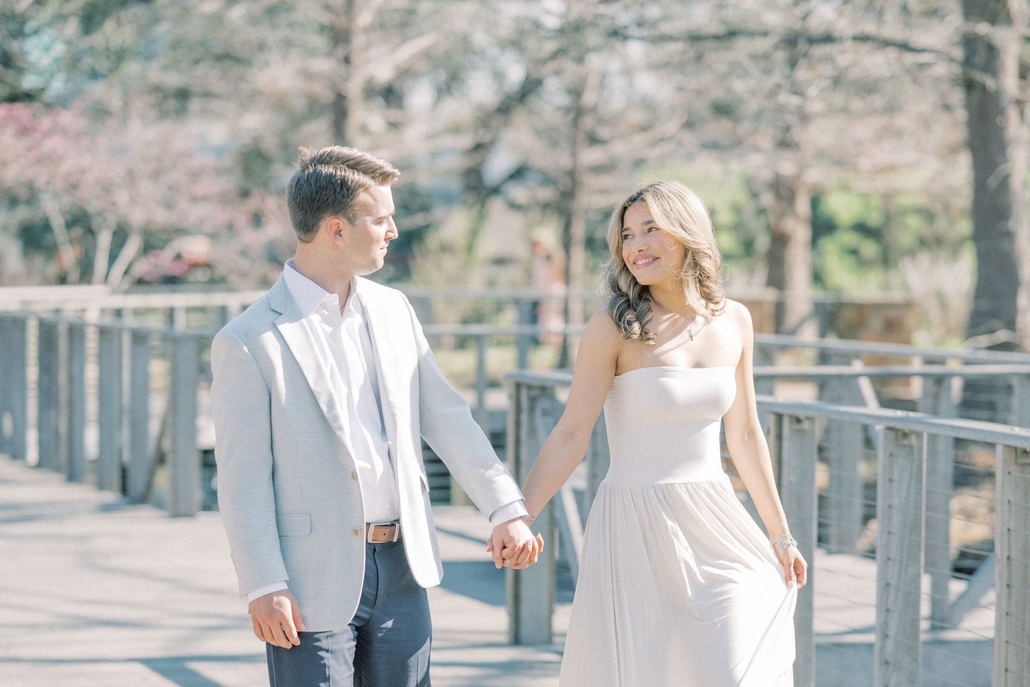 A couple in elegant wedding attire walks along a wooden boardwalk surrounded by cherry blossoms in spring.