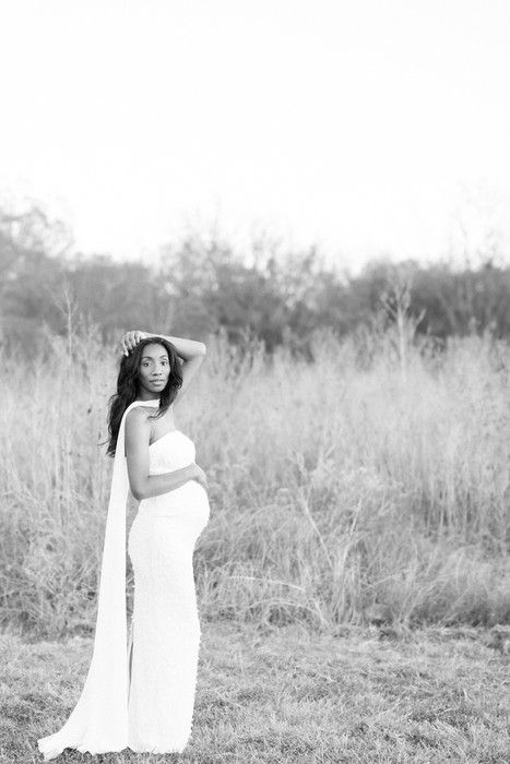 Black and white maternity photo of an expectant mother in a flowing white dress standing in a sunlit field.