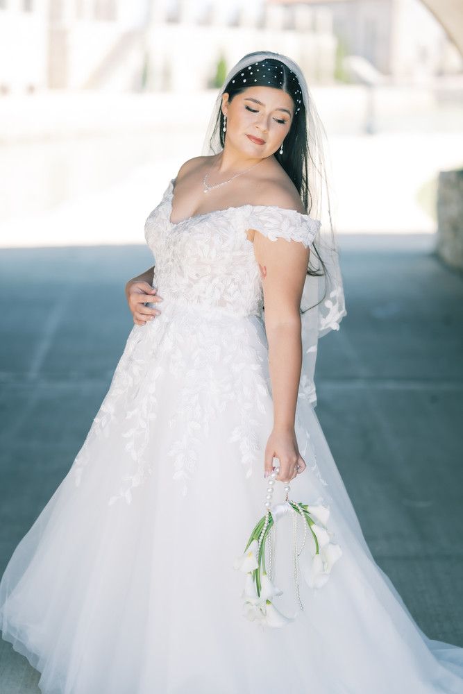 A bride in an elegant off-shoulder white lace wedding gown holds white calla lilies while posing in soft lighting.