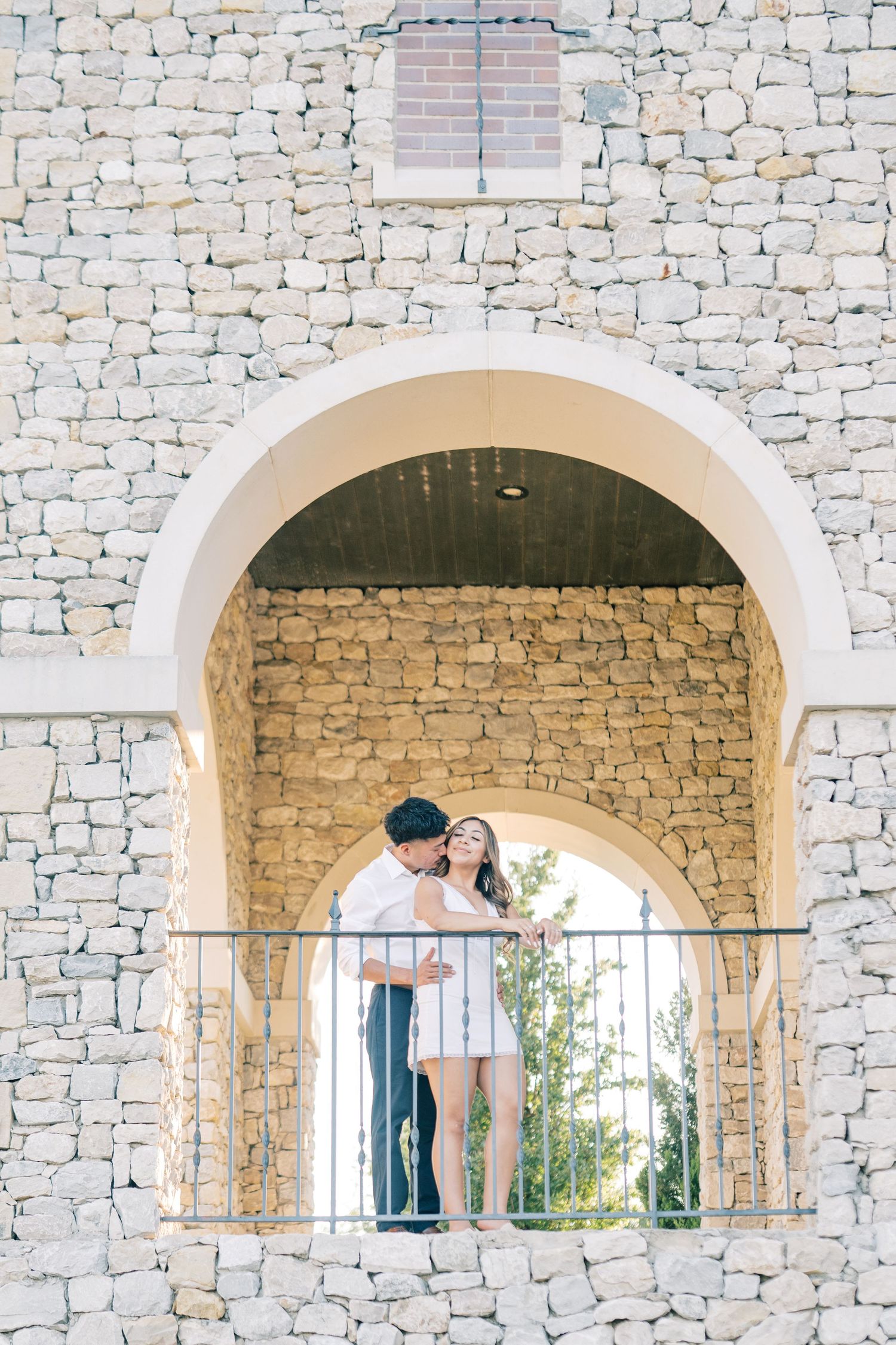 Couple sharing a tender moment under a grand stone arch on a sunlit balcony.