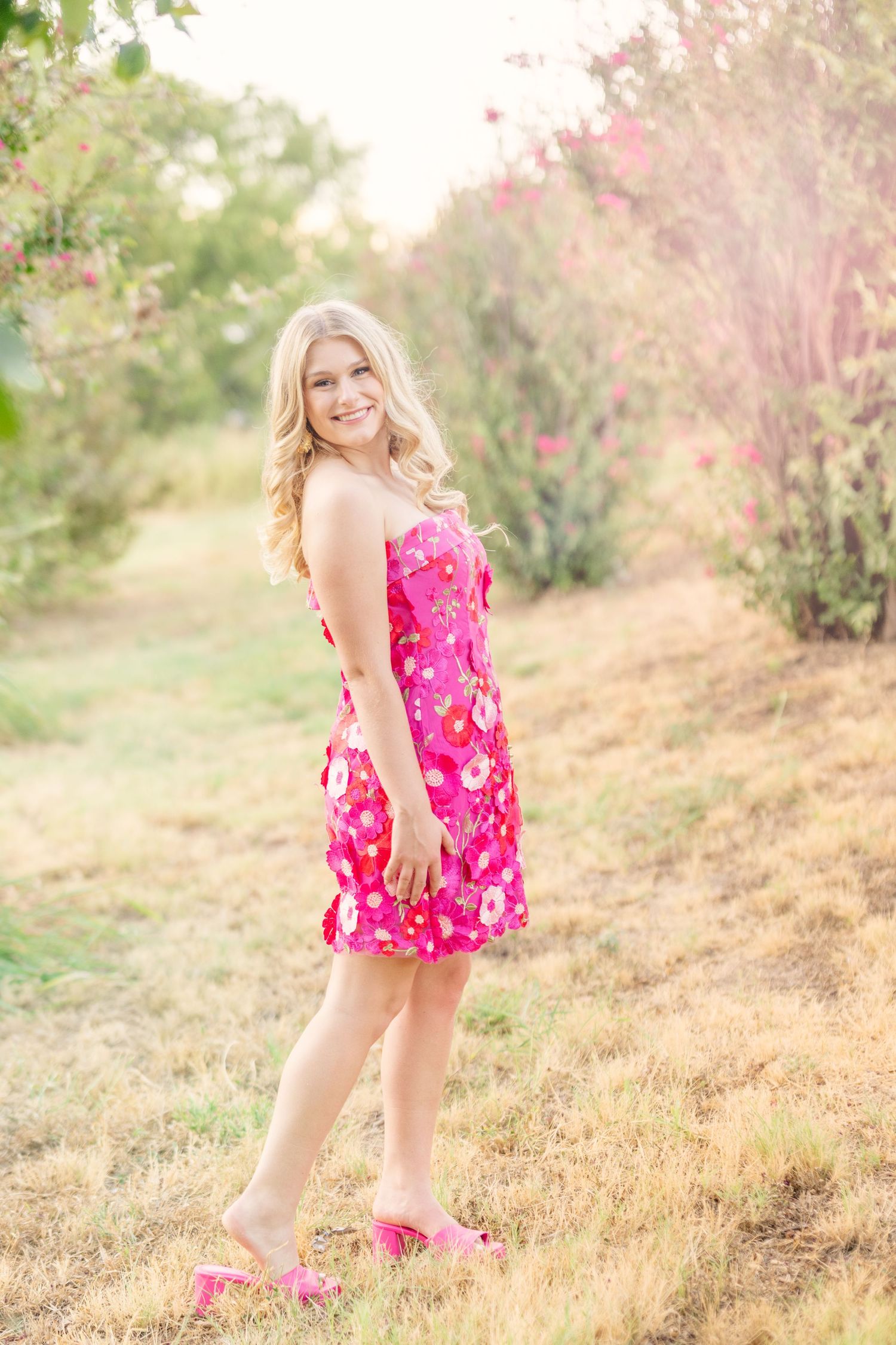 Smiling blonde woman in pink floral sundress and pink heels posing outdoors among blooming trees.