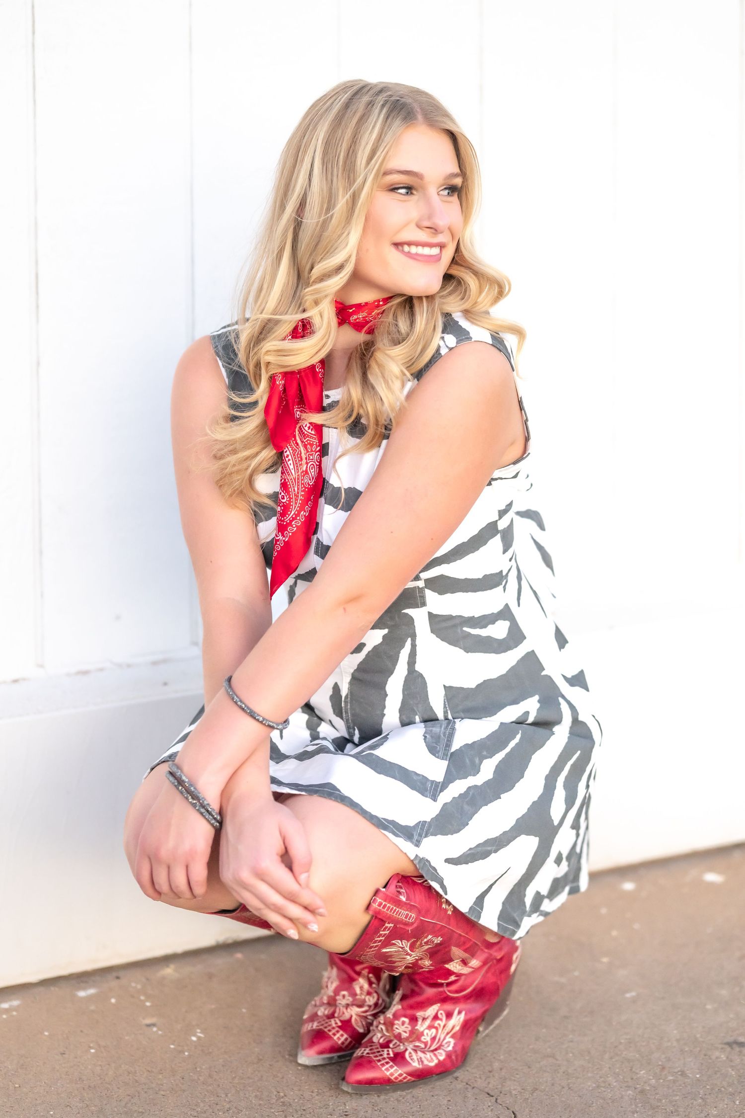 Smiling blonde woman in zebra print dress, red bandana, and red cowboy boots crouching against a white wall.