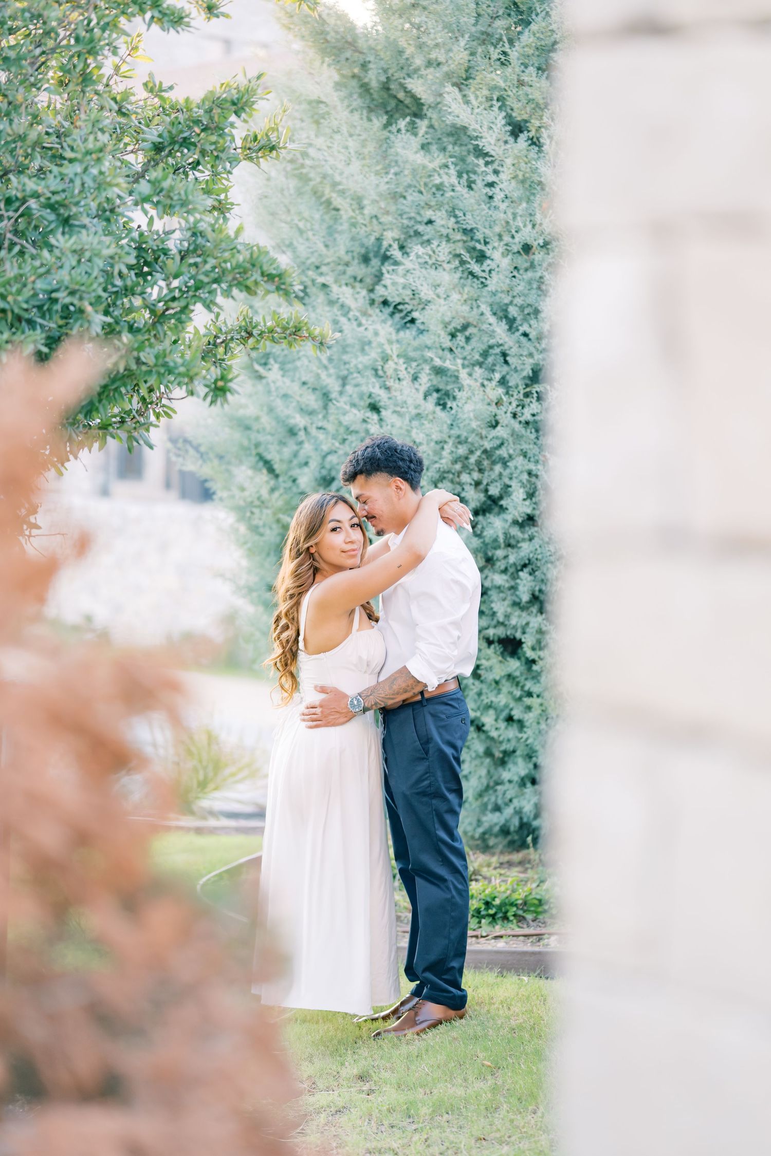 A couple shares a romantic embrace surrounded by lush green trees during their outdoor engagement shoot.