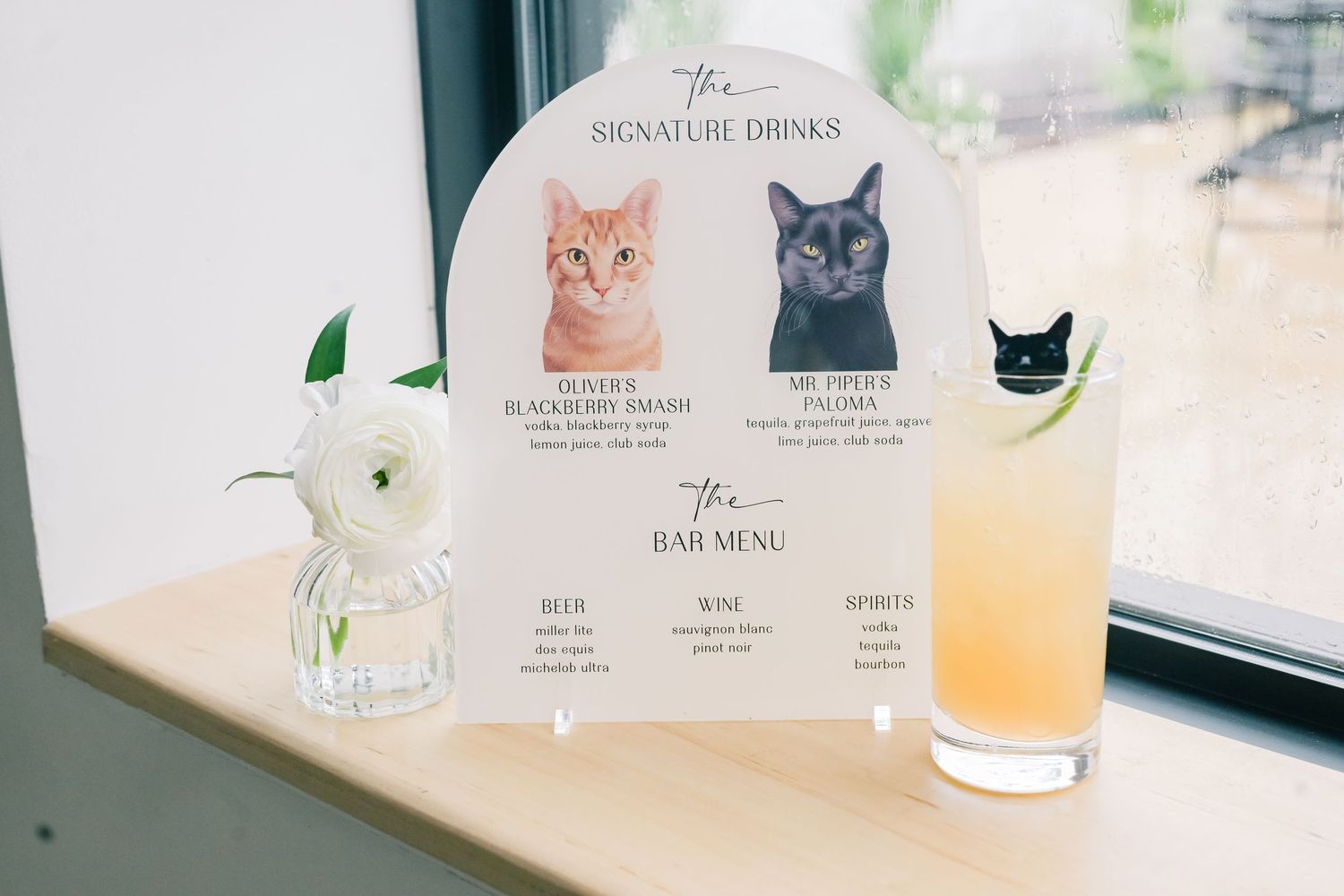 Arched wedding signature drinks menu sign featuring illustrated cats beside a cocktail and white flower.