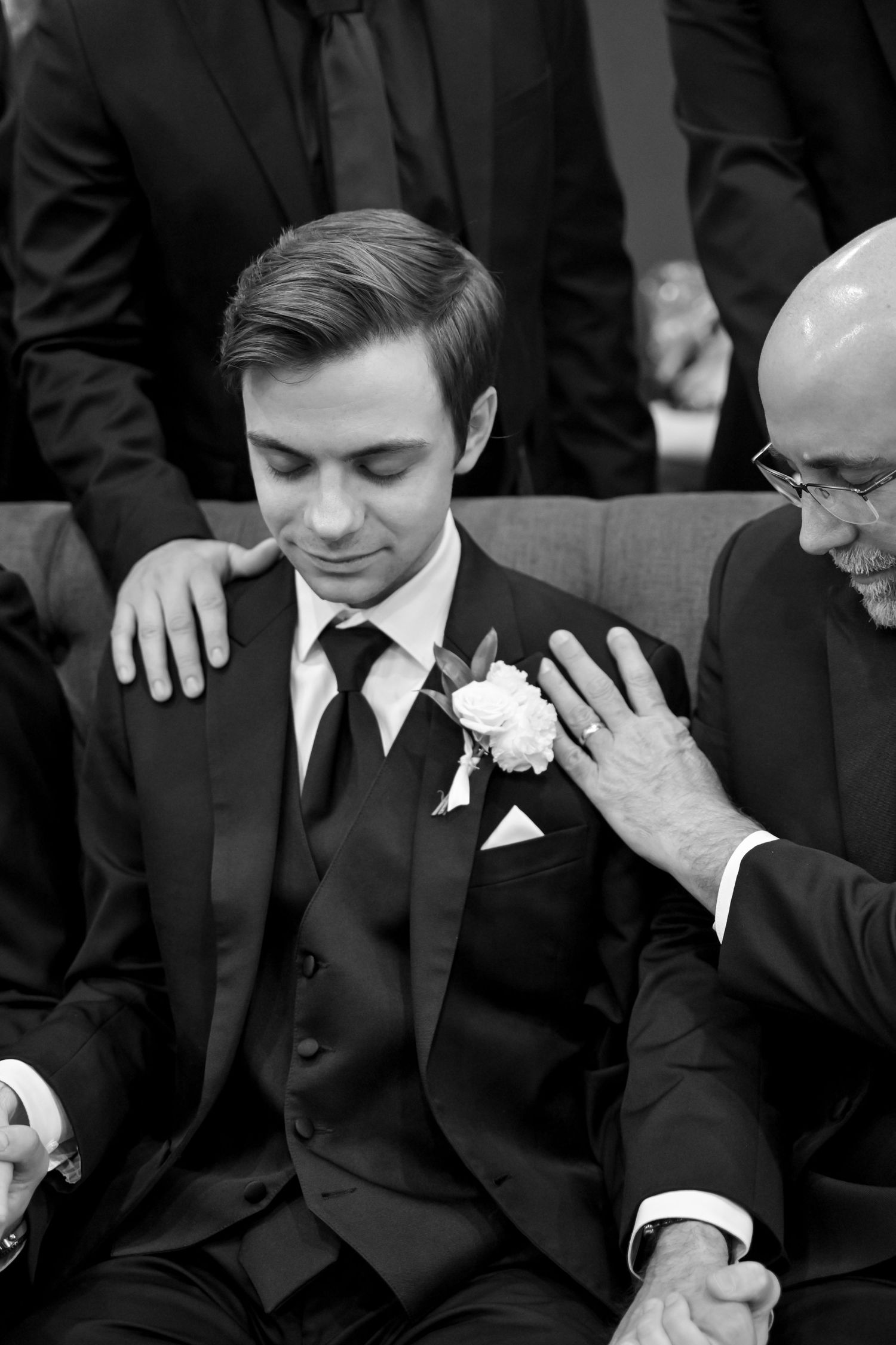 Groom in a three-piece suit with boutonniere receives comforting gestures from loved ones during wedding ceremony.