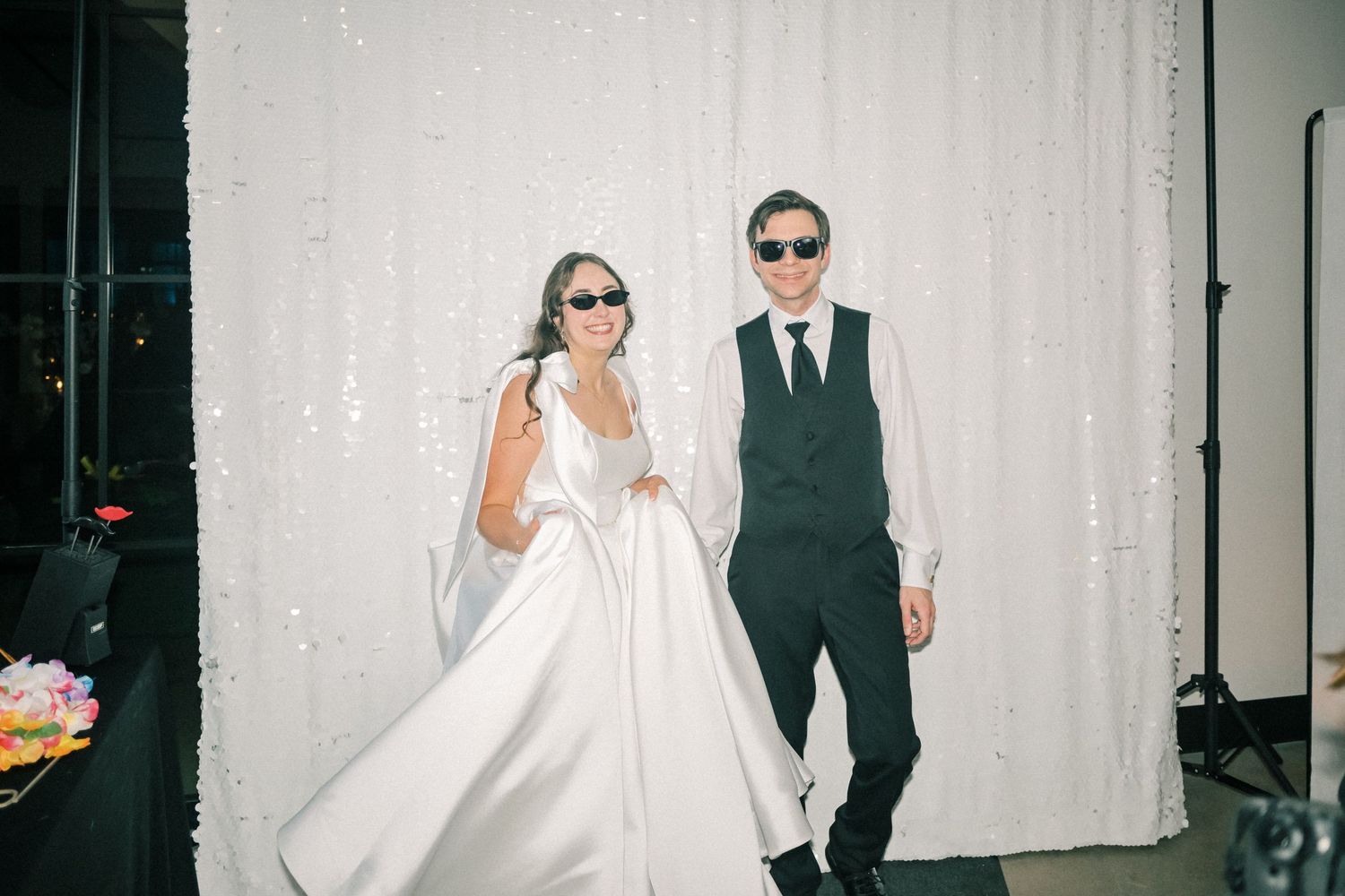 Bride and groom wearing sunglasses posing together in front of a white sequin backdrop at their wedding reception.