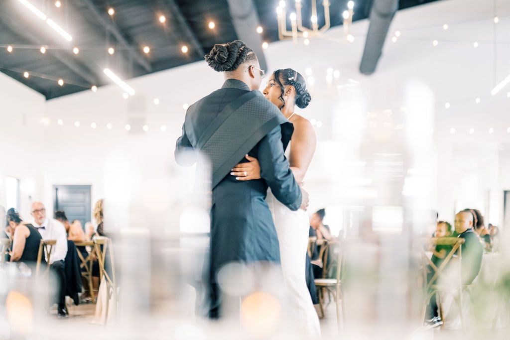 Bride and groom share their first dance at elegant wedding reception with string lights and guests seated in background.