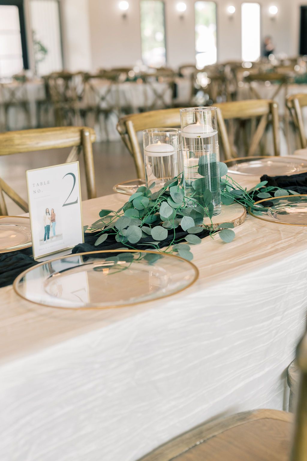 Wedding reception table with eucalyptus greenery, floating candles, gold charger plates, and table number card.