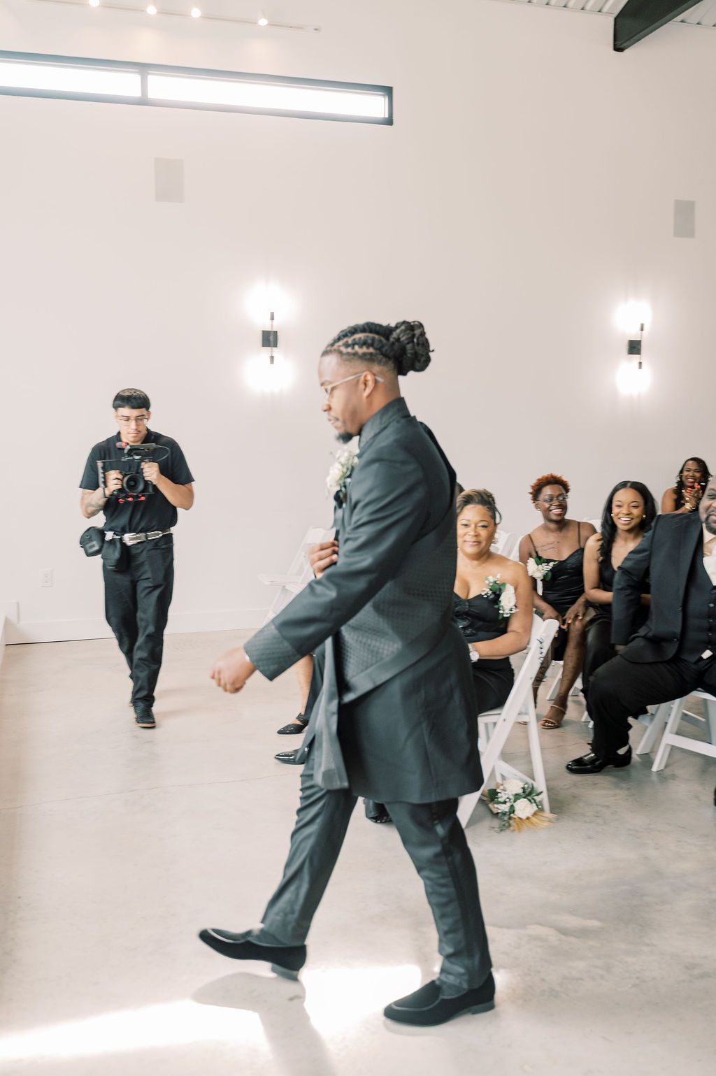 Groom walks down the aisle in a dark suit at a modern wedding venue with seated guests.