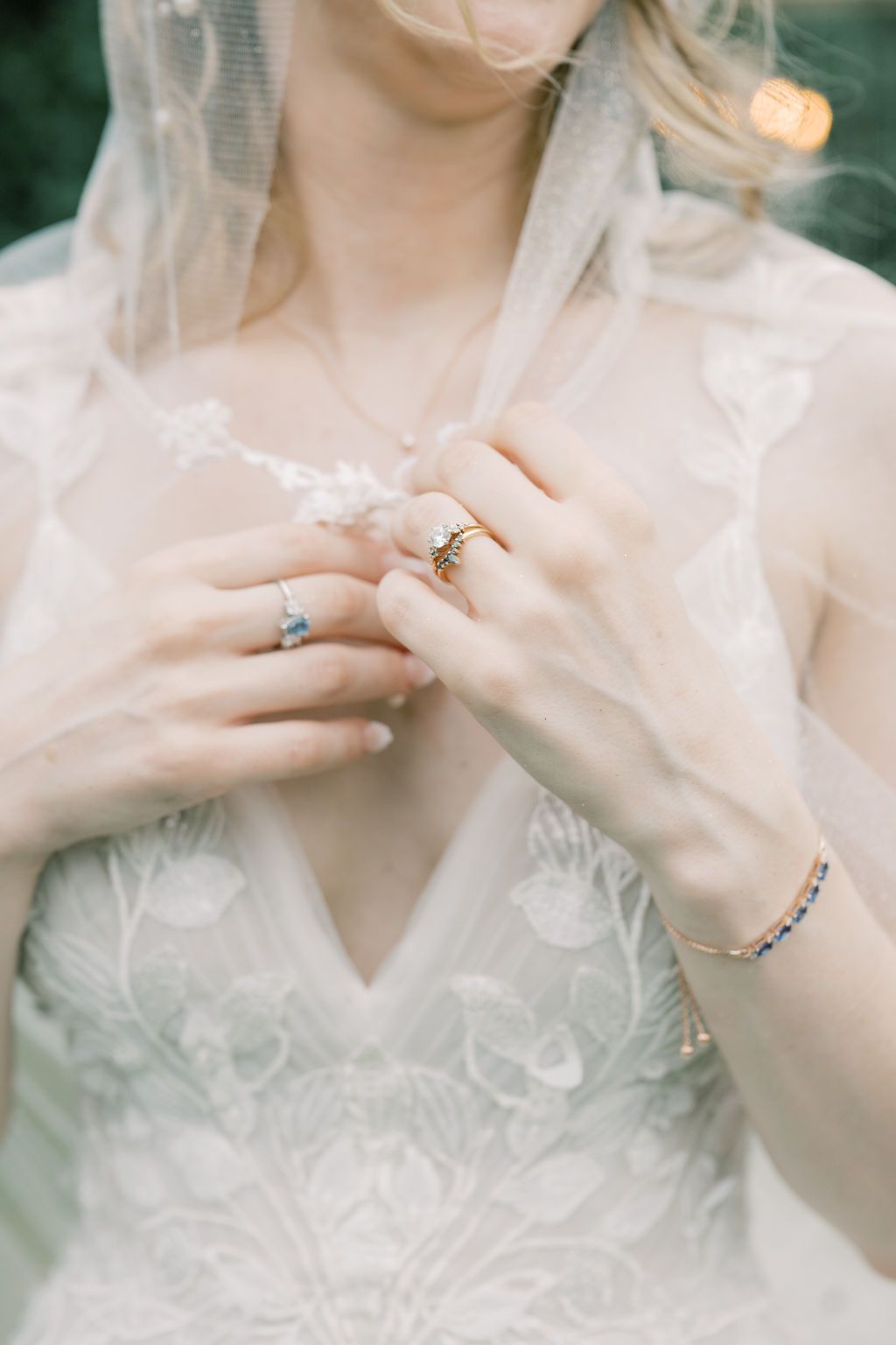Bride holding veil wearing gold engagement ring, sapphire band, and delicate bracelet over lace wedding dress.