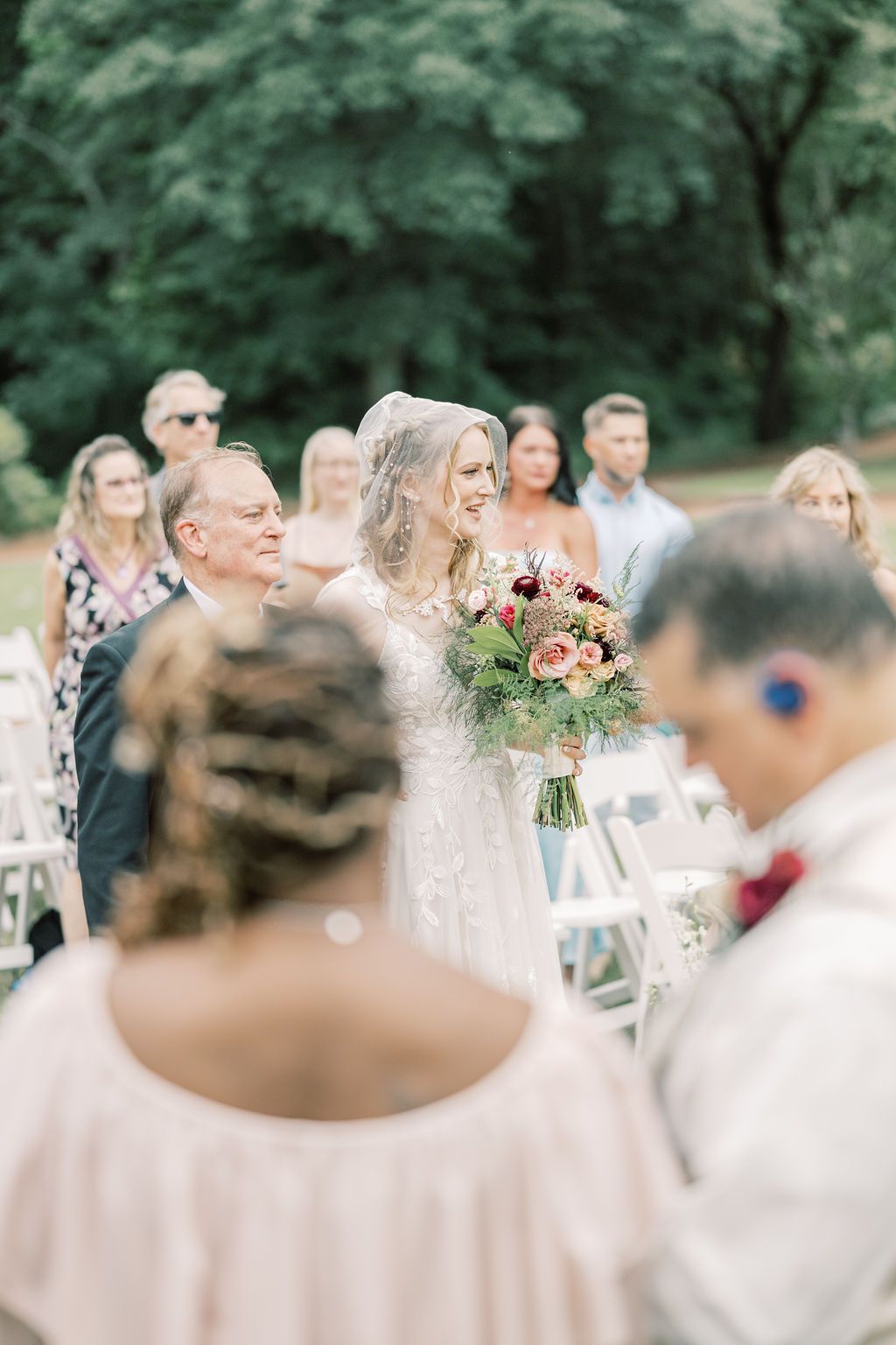 Bride walking down the aisle holding a lush floral bouquet, smiling at guests during an outdoor wedding ceremony.
