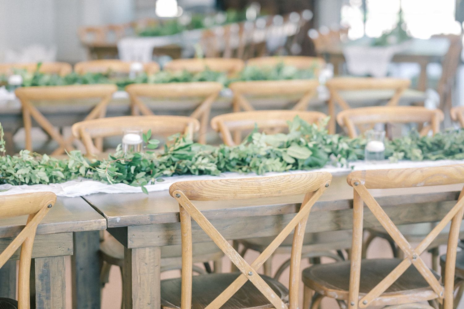 Rustic wooden cross-back chairs at reception tables adorned with lush green leaf garlands.
