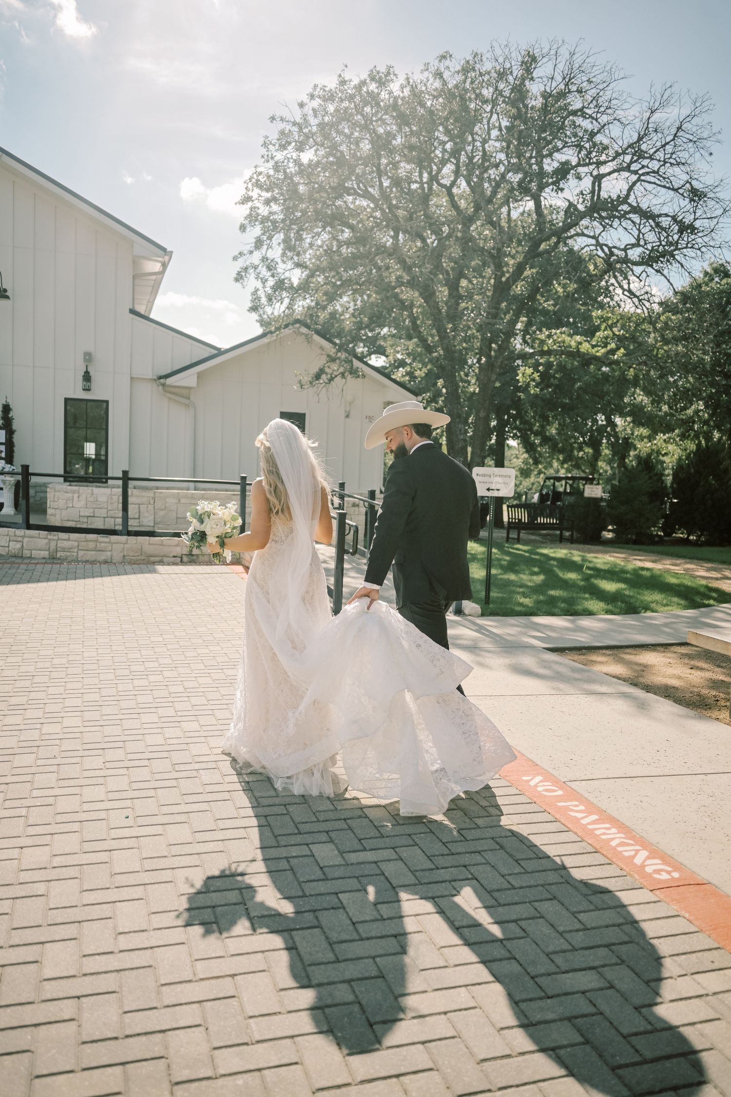 Bride and groom walking hand in hand on brick path toward white barn wedding venue in sunlight.