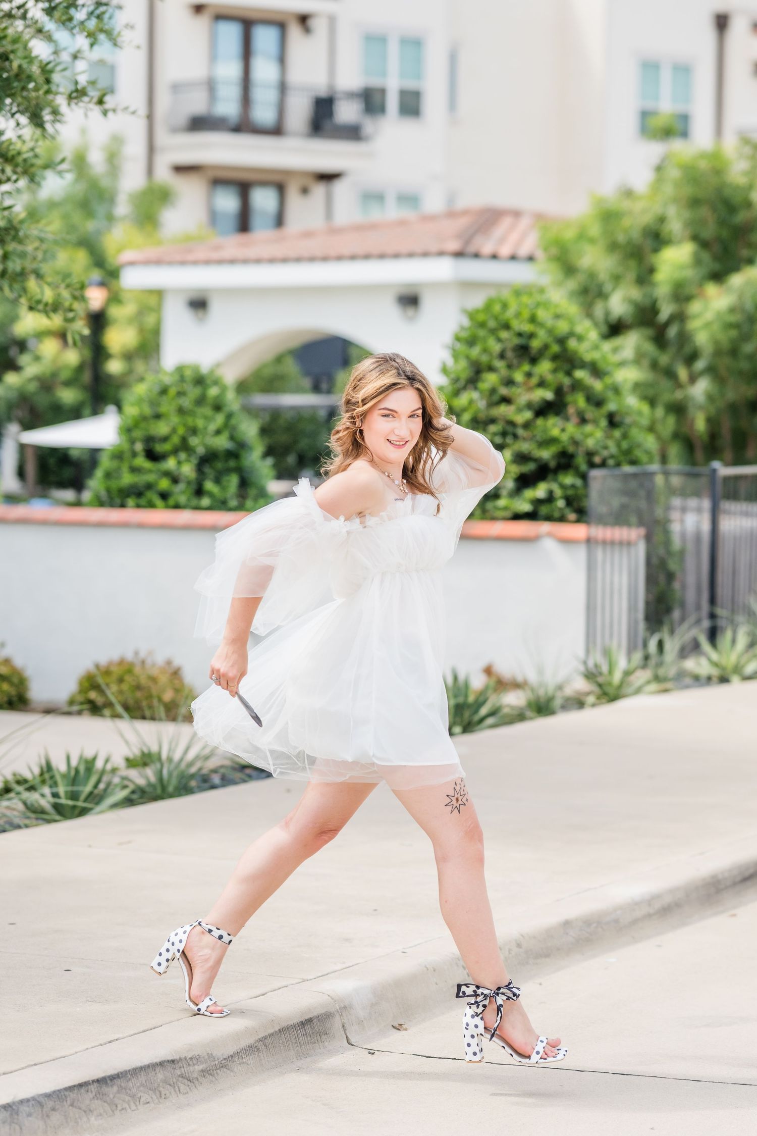 Young woman in white off-shoulder mini dress with polka dot heels posing outdoors near Mediterranean-style building.