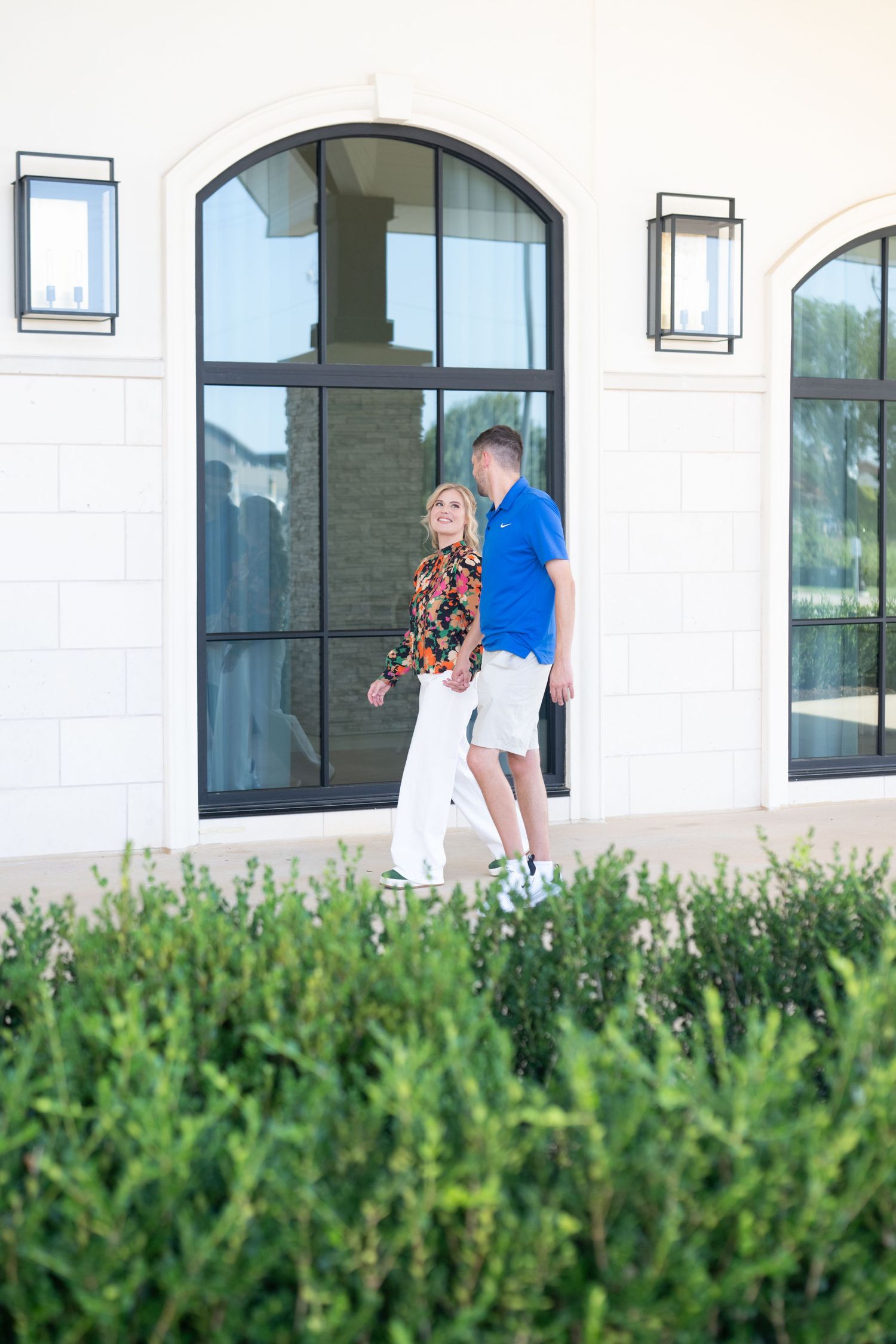 Couple walking hand in hand past arched windows on a white building exterior.