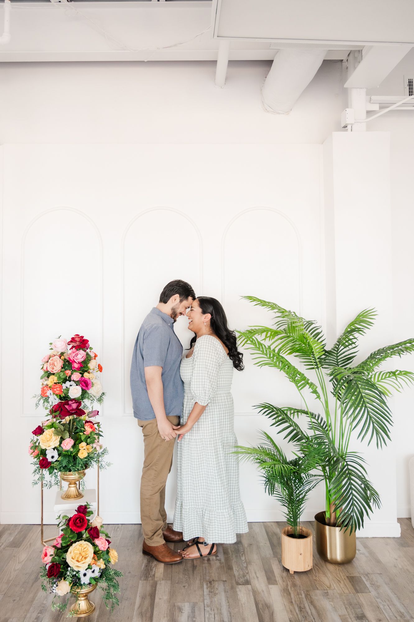 Romantic couple sharing an intimate moment against white wall with floral arrangements and palm plants.