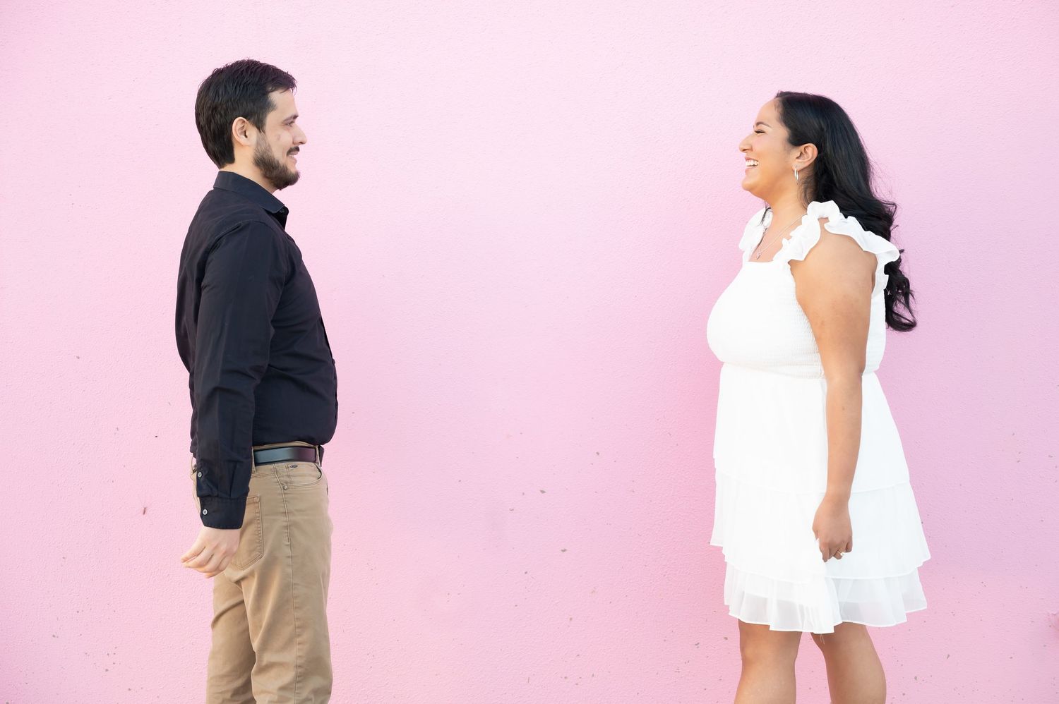 Couple in contrasting white dress and black shirt pose against pink wall in romantic photoshoot.
