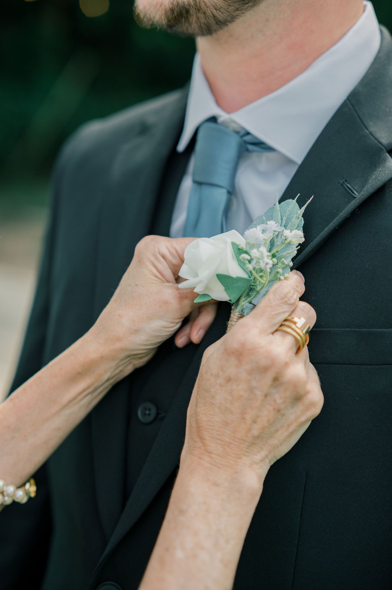 Hands pinning a light blue and white floral boutonniere onto a dark suit lapel.