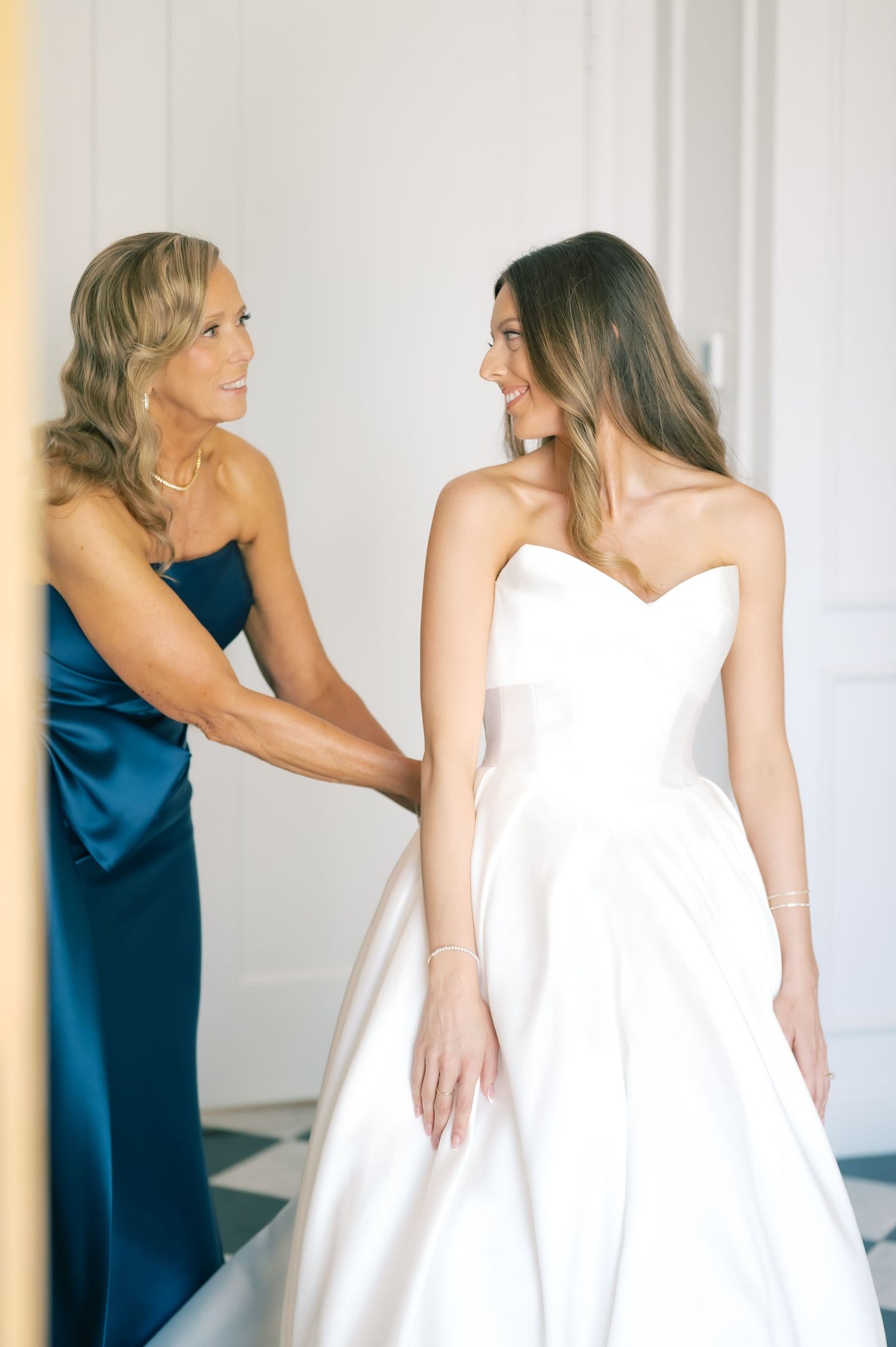 Bride in strapless white gown smiles at woman in blue dress helping her prepare for wedding day.