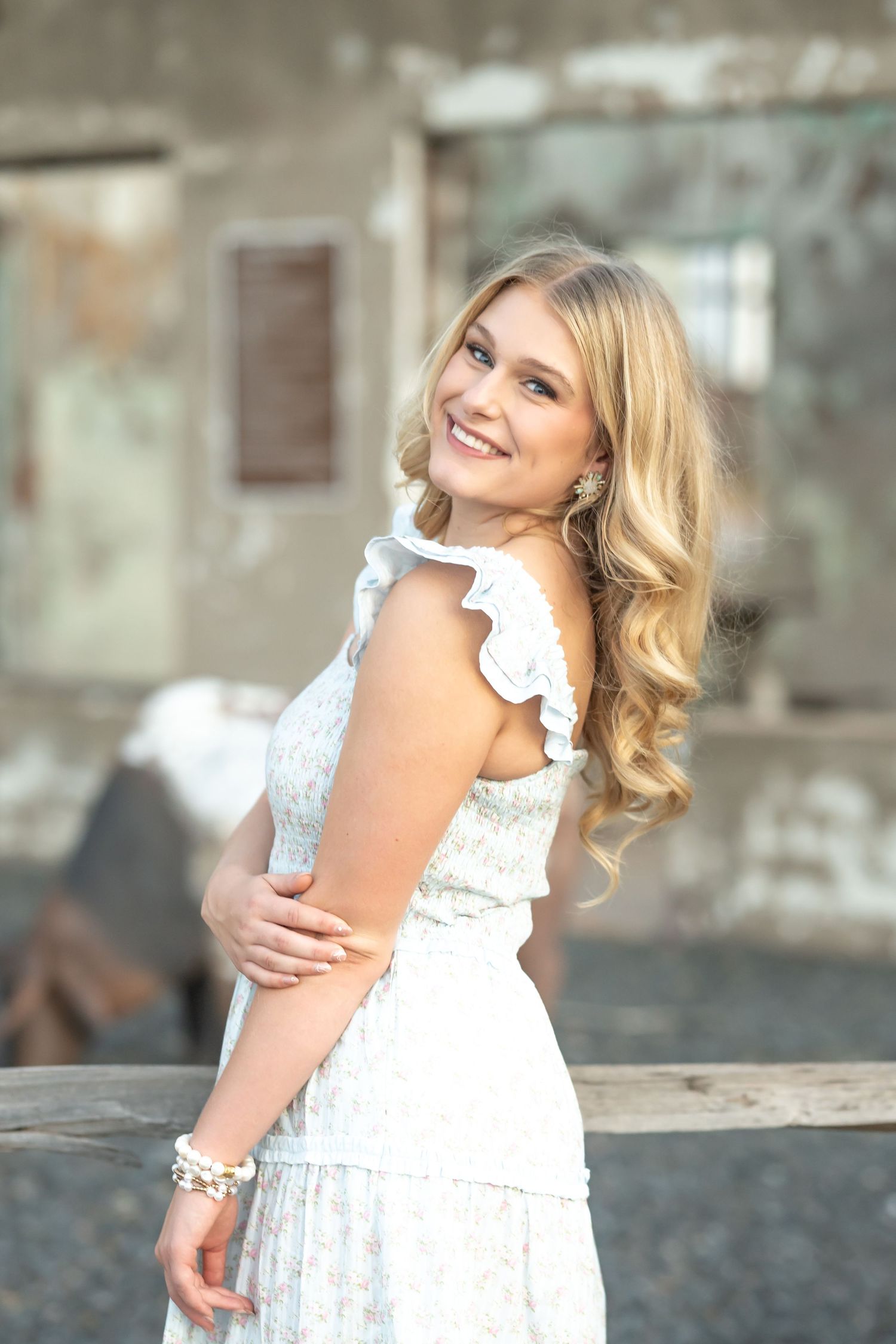 Smiling blonde woman in a white floral ruffled dress posing outdoors near a rustic wooden fence.