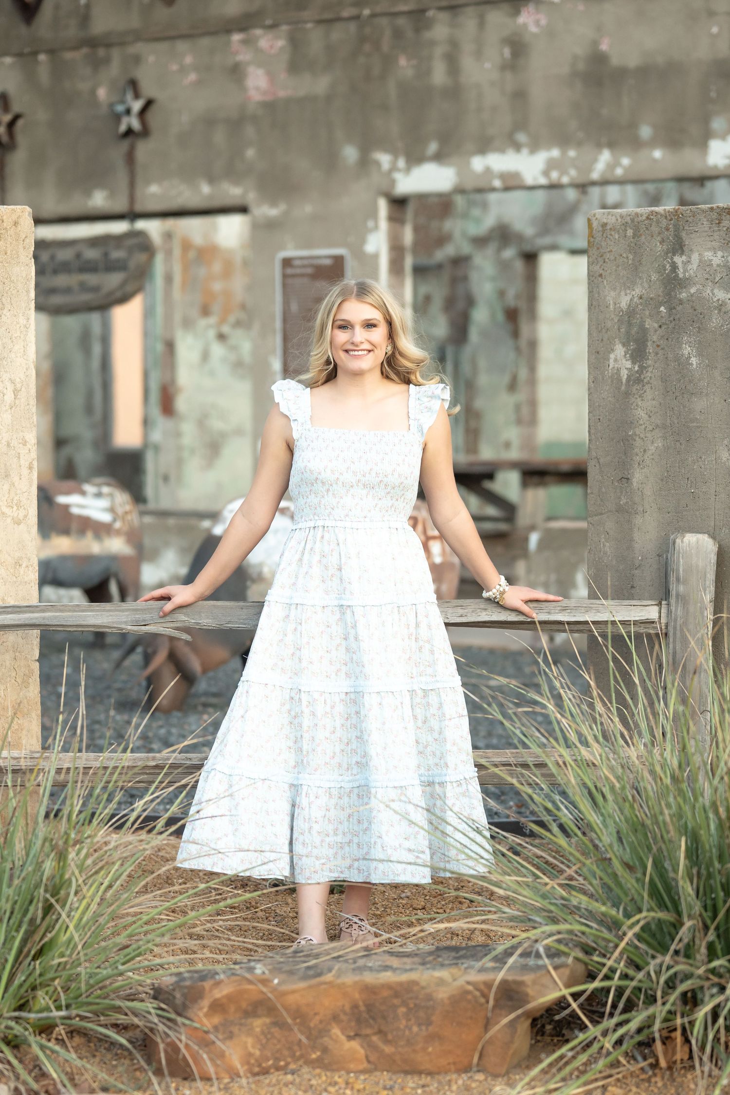 Young blonde woman in a white floral midi dress poses outdoors near rustic wooden fence and weathered building.