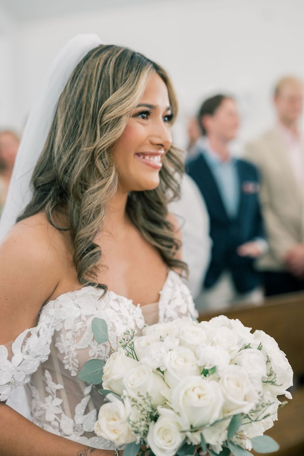 Smiling bride in lace off-shoulder gown holds white rose bouquet with eucalyptus at church wedding ceremony.
