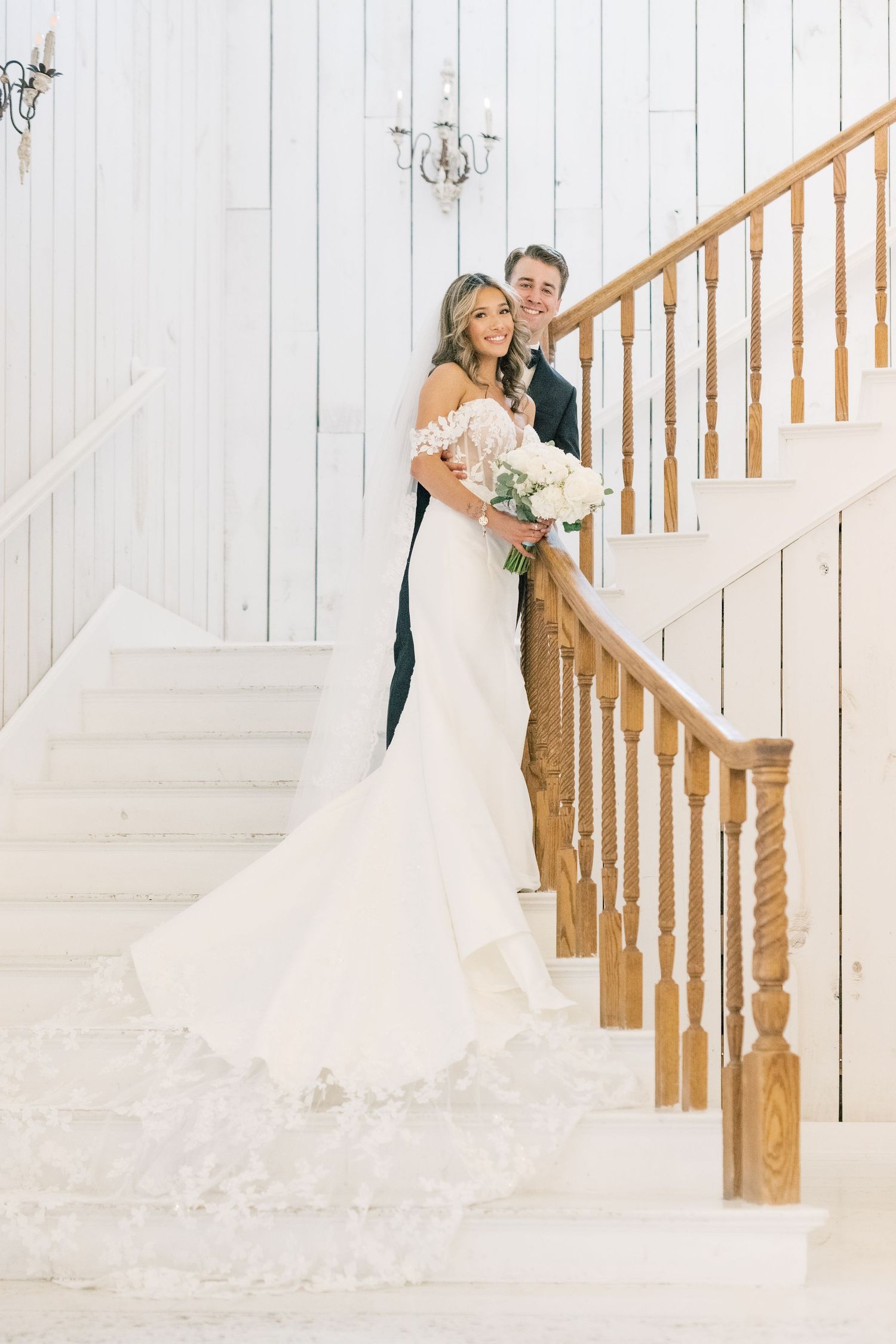 Bride and groom posing on white staircase with wooden railing, bride holding white floral bouquet.