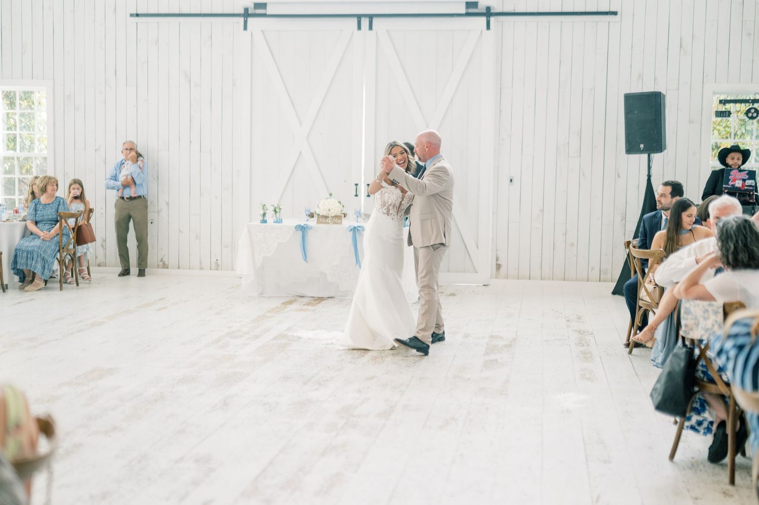 Bride and groom sharing first dance in rustic white barn reception hall surrounded by seated wedding guests.