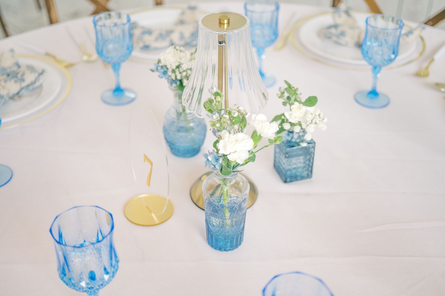 Elegant wedding table setting with blue glassware, white roses, and gold accents on a white tablecloth.