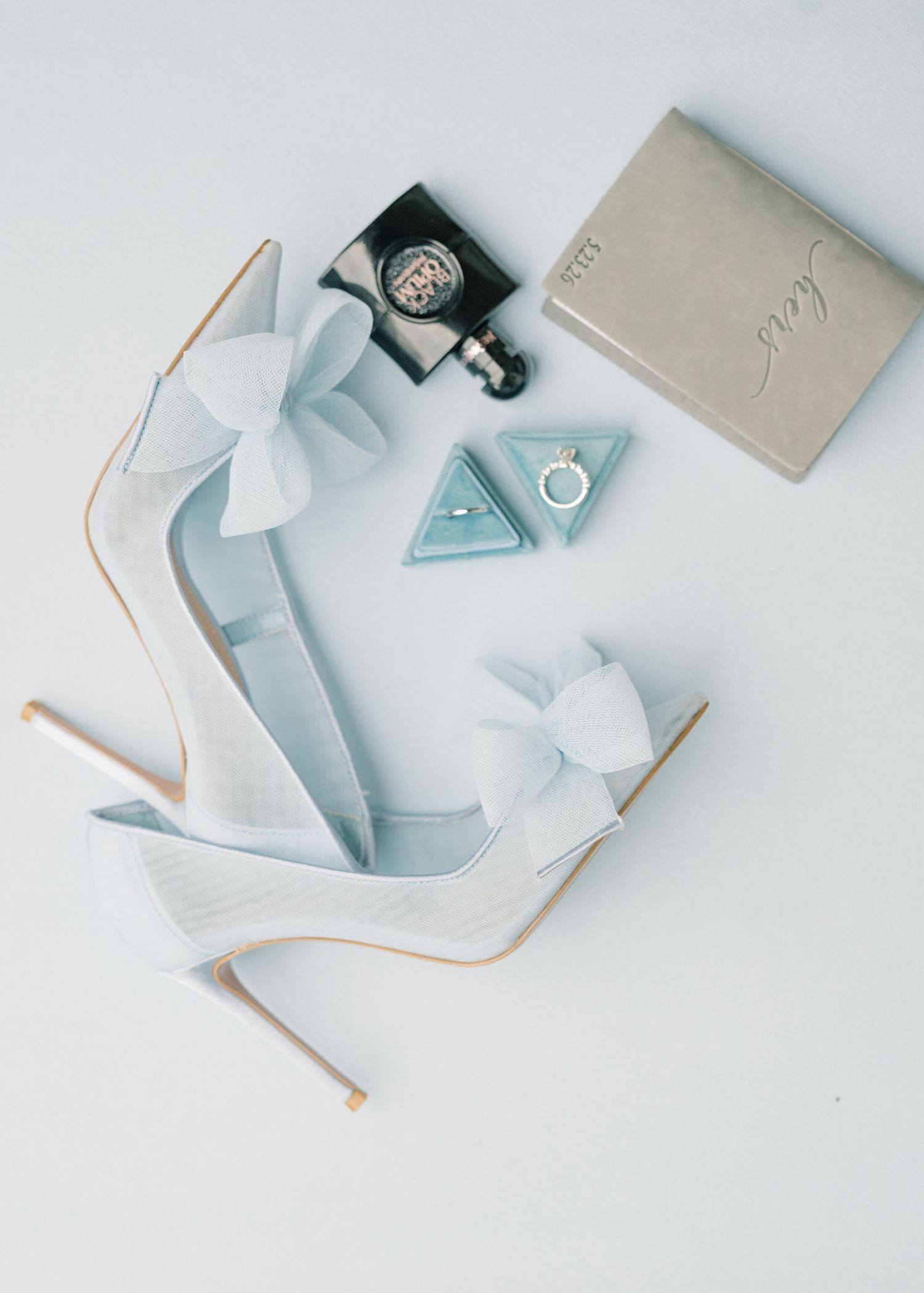 Light blue bridal heels with bow detail beside wedding rings and perfume bottle on white background.