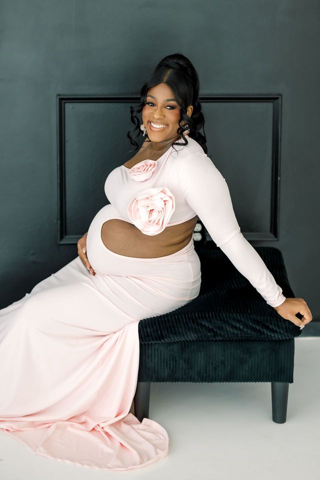 Pregnant woman in pink cutout maternity gown with floral detail seated gracefully on dark velvet bench.