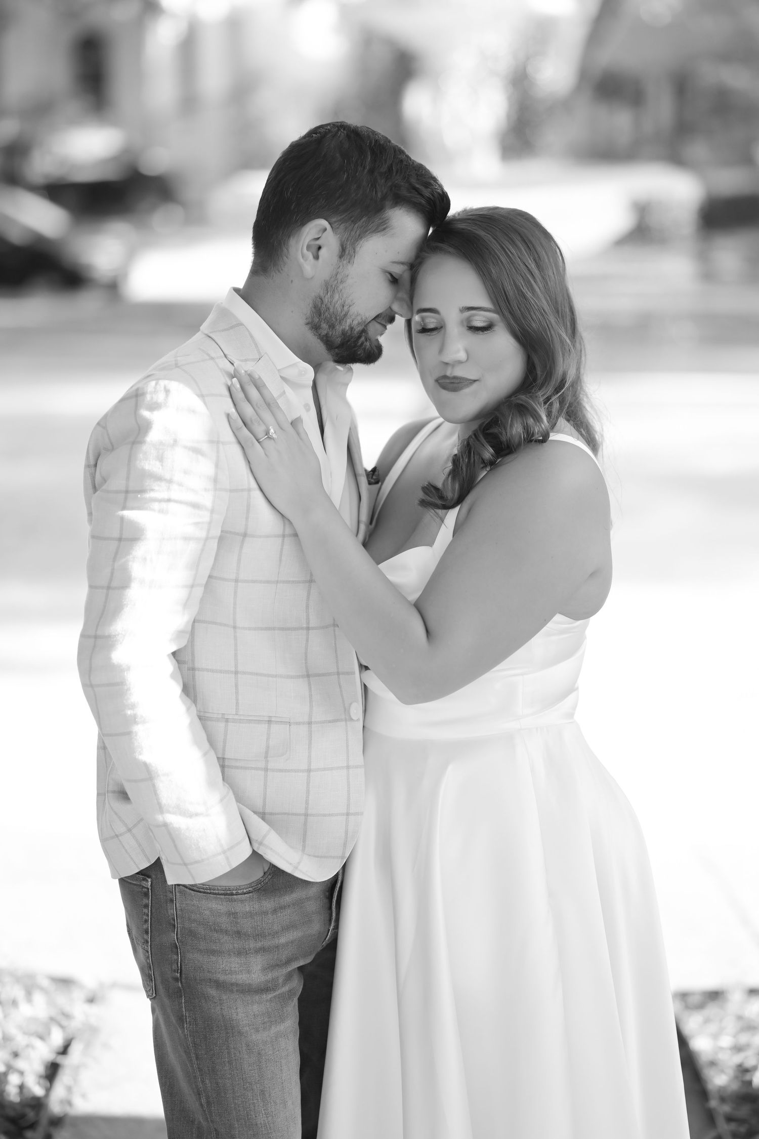 Black and white engagement portrait of a couple embracing with foreheads touching, the woman in a white dress with eyes closed.
