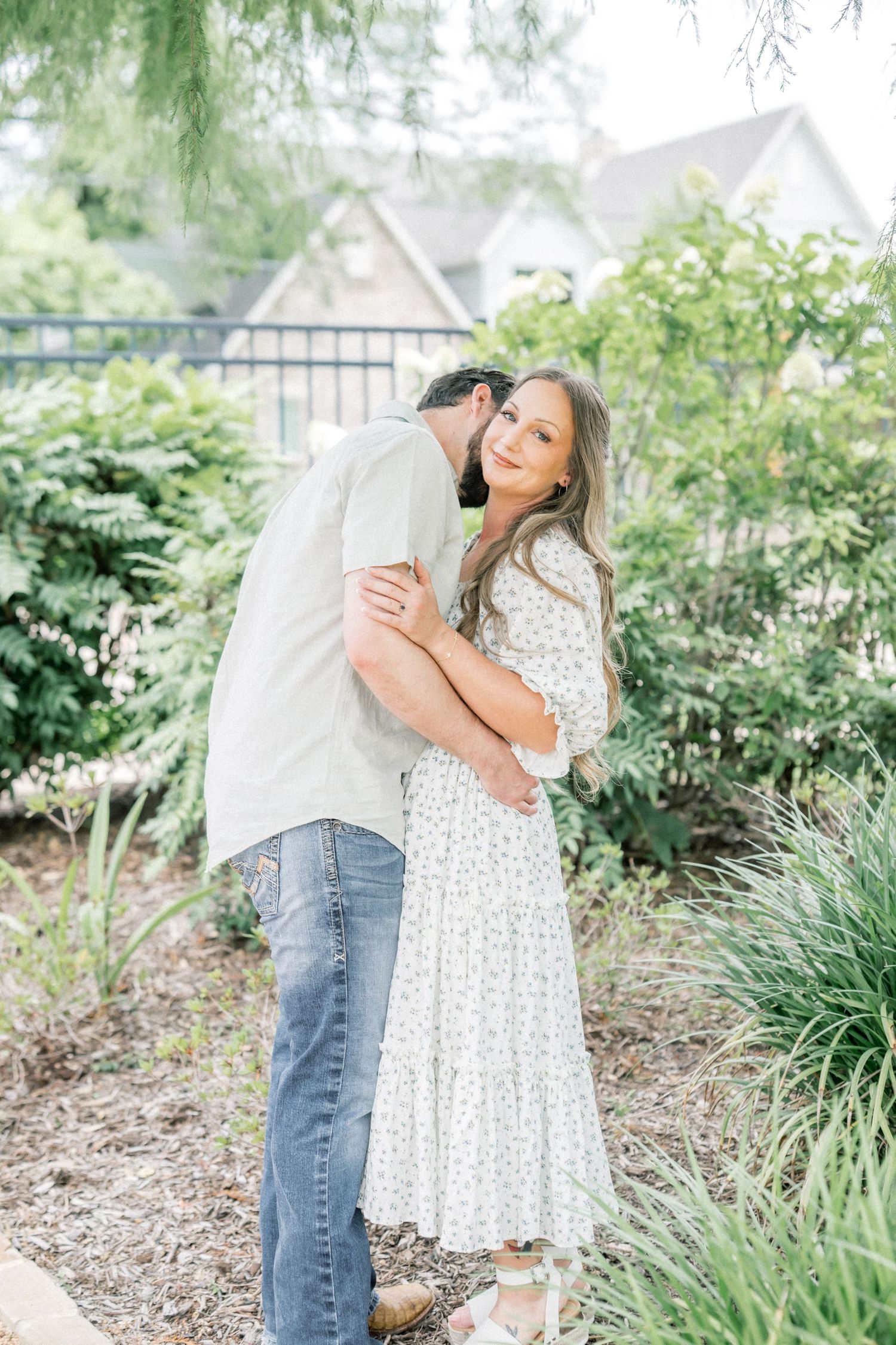 Man kissing woman on cheek as she smiles in a lush garden setting, both dressed casually.