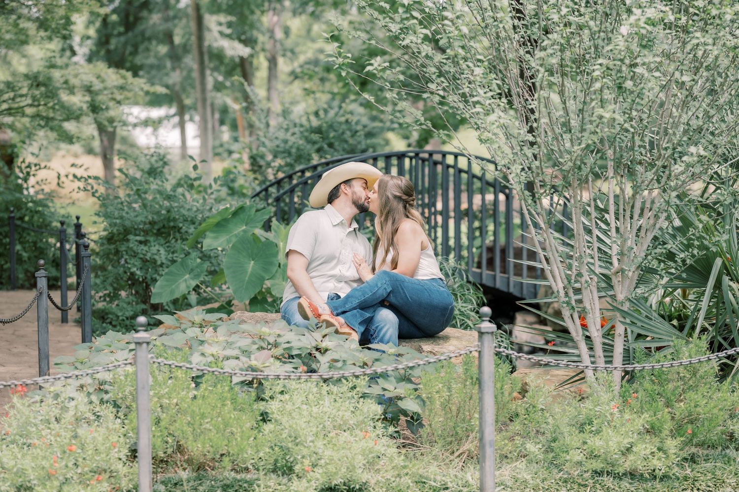 Couple sharing a kiss while sitting outdoors near a decorative bridge surrounded by greenery.