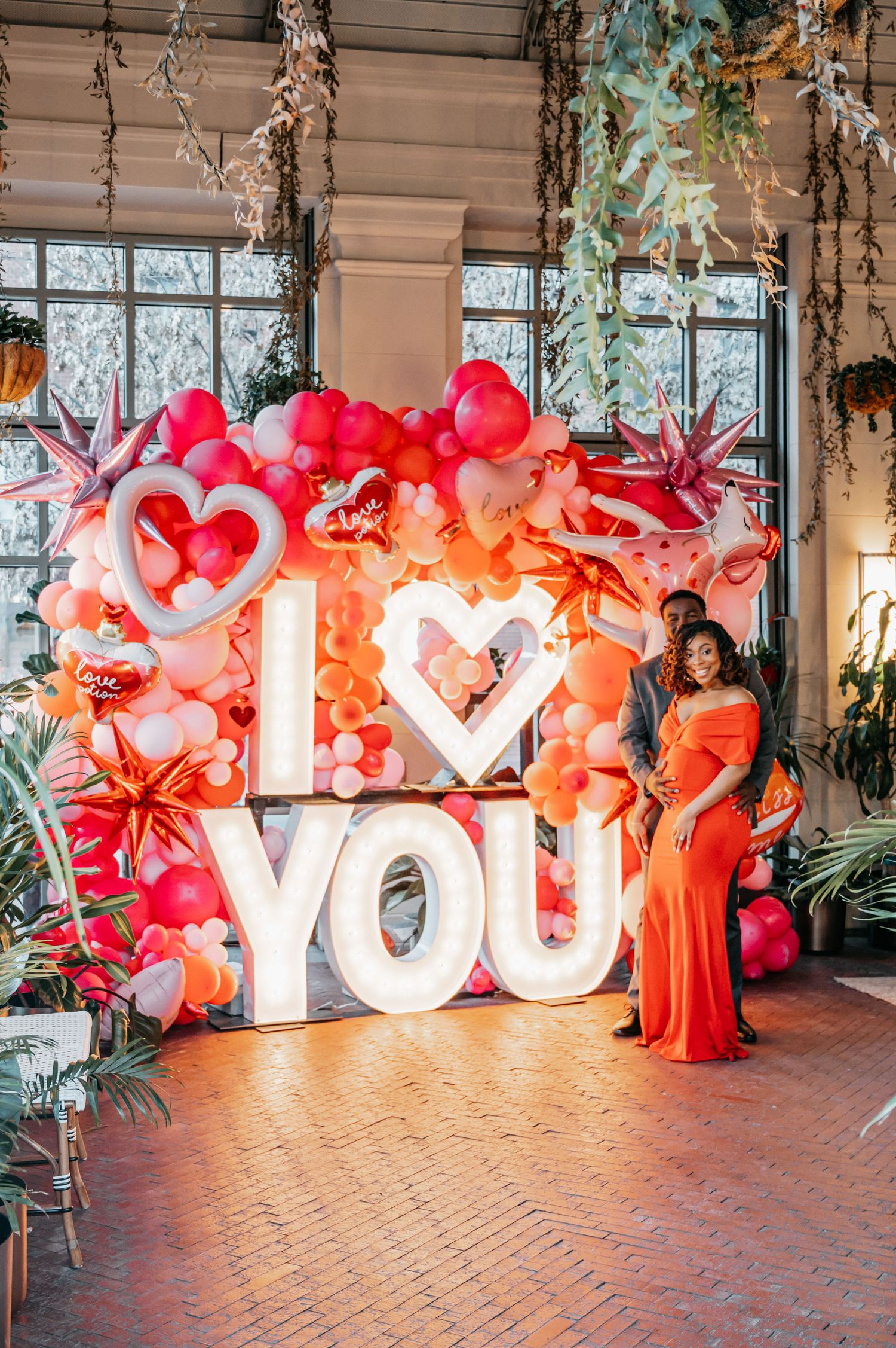 Couple poses before illuminated I Love You sign surrounded by pink and red balloon garland in elegant venue.