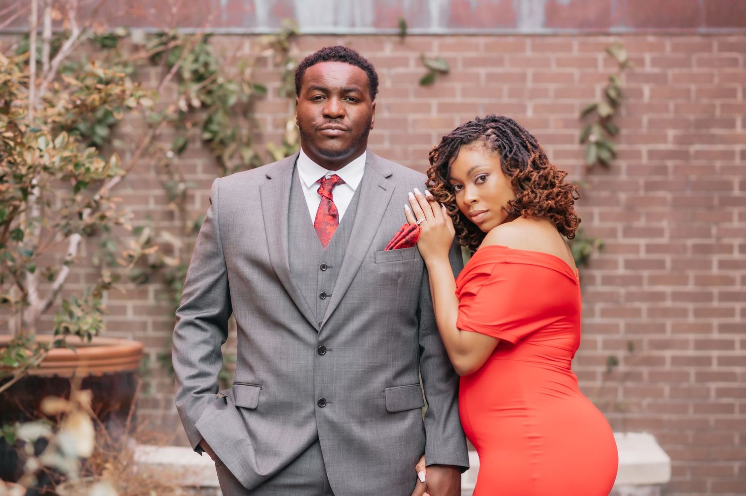 A man in a gray suit and a woman in a red dress pose together outdoors near a brick wall.