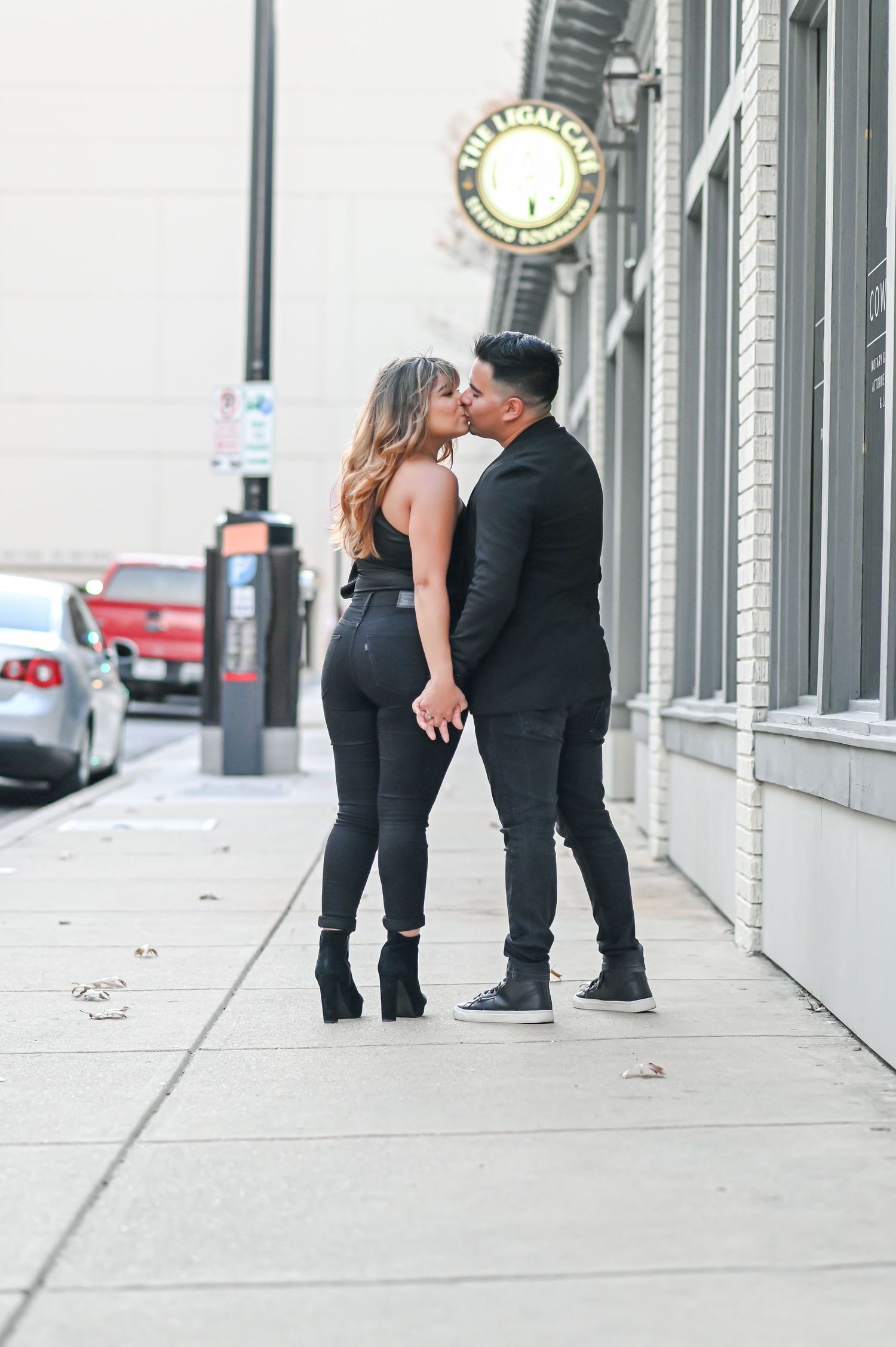 Couple sharing a kiss on an urban sidewalk wearing coordinated black outfits.