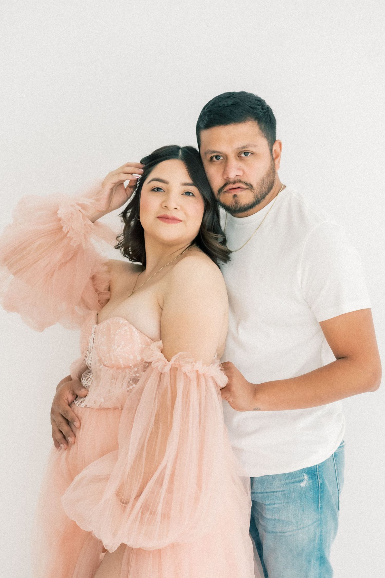 Pregnant woman in pink tulle gown poses with partner in white t-shirt against white background.