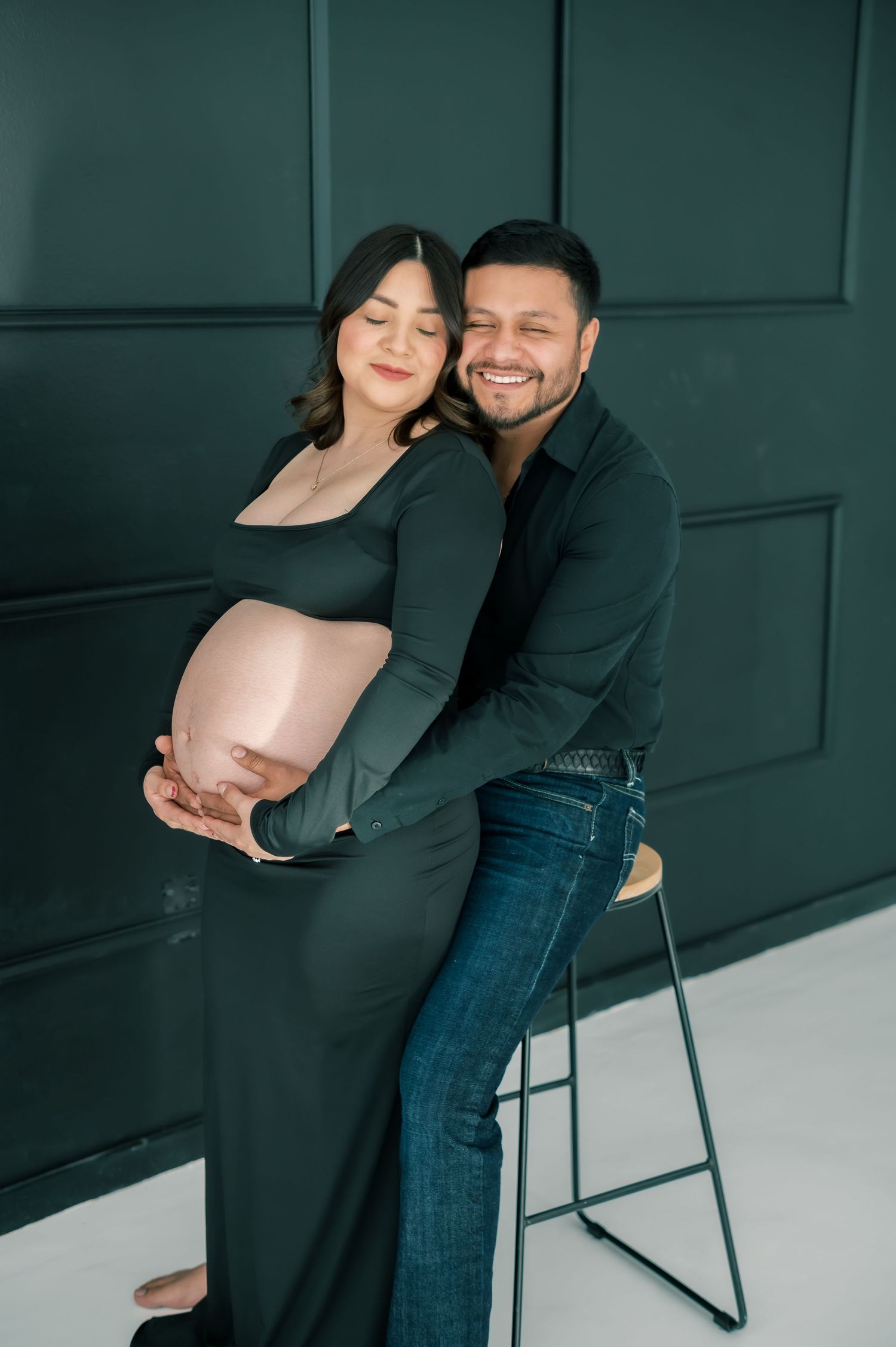A smiling couple in a maternity photo shoot, man embracing pregnant woman in dark green dress against teal paneled wall.