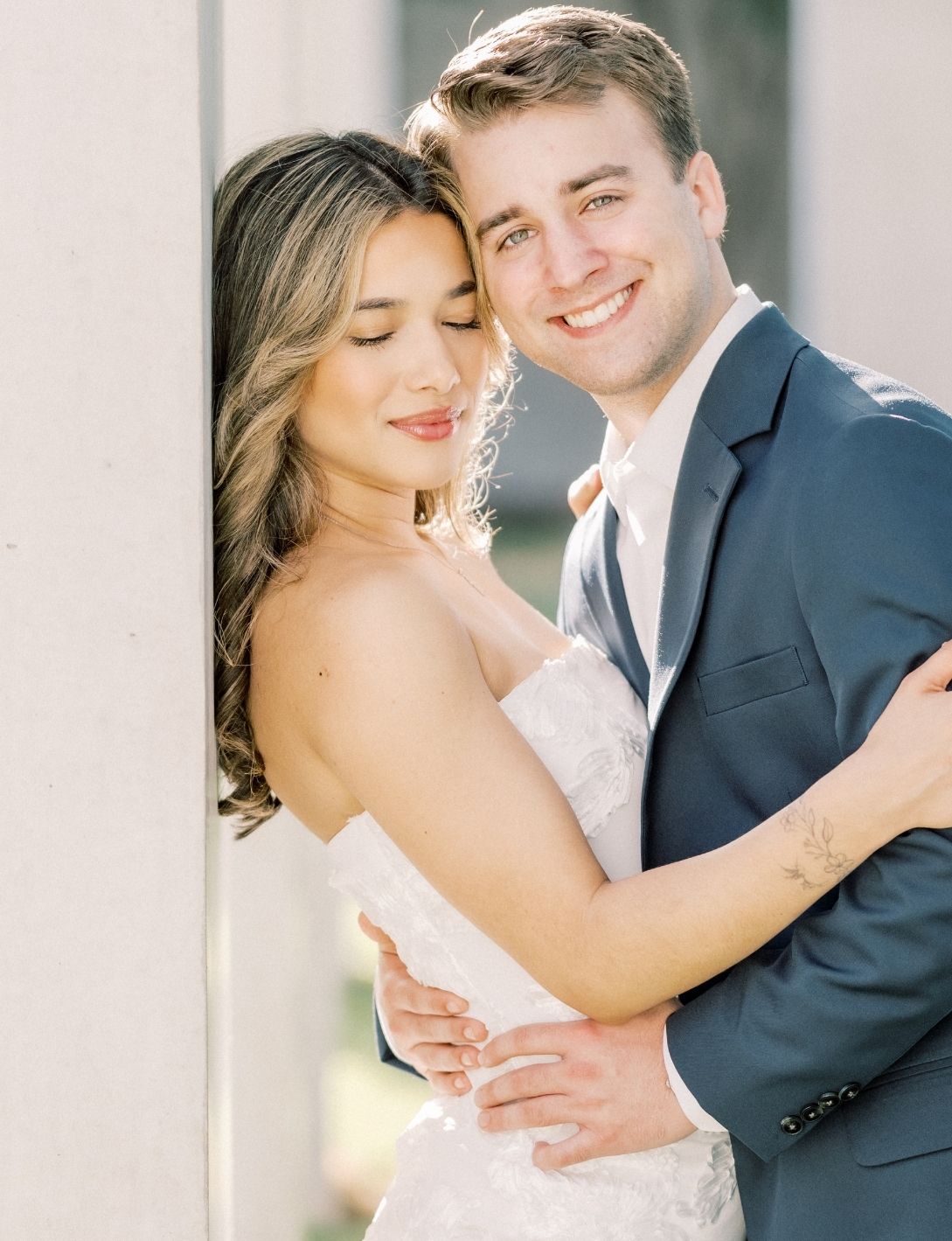 Couple embracing in wedding attire, bride in strapless lace gown and groom in navy suit, posing against a column.