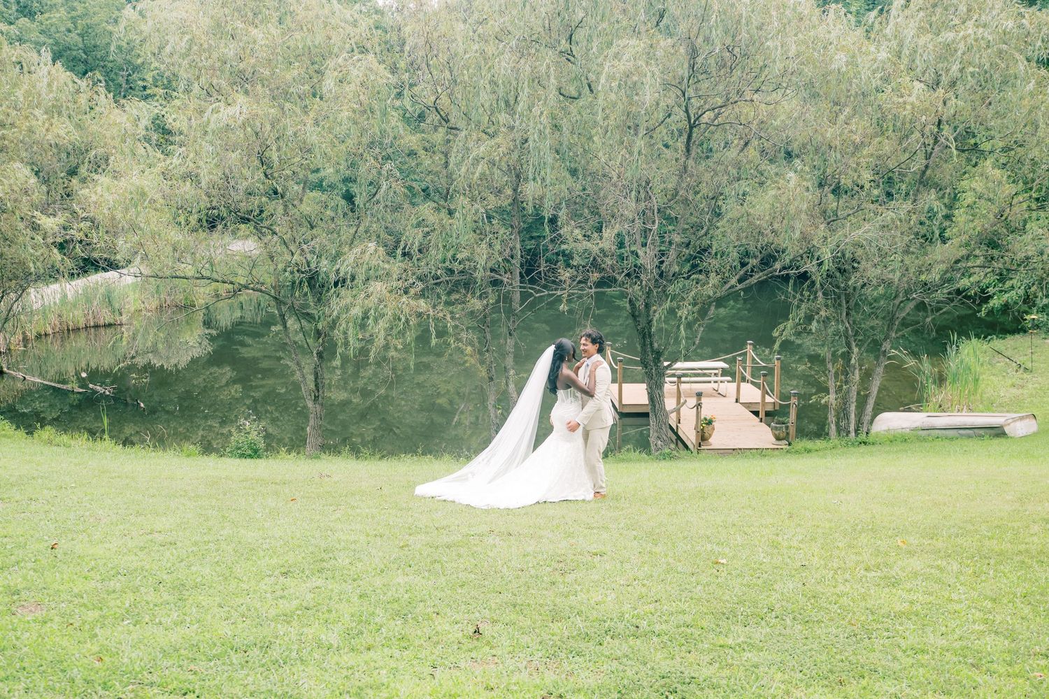 Bride and groom share romantic kiss in lush green meadow with trees and wooden structure in background.