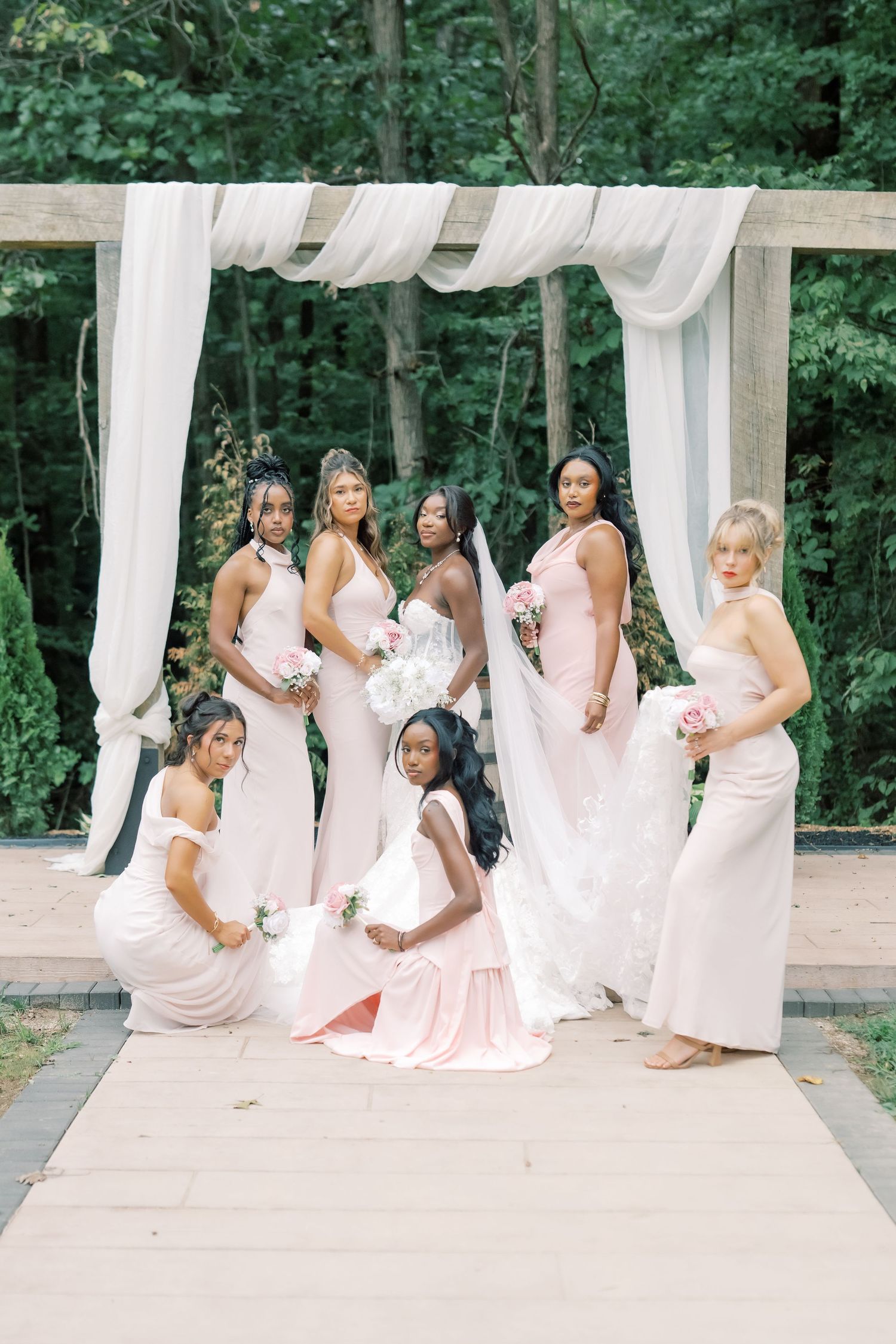 Bride in lace gown with bridesmaids in blush pink dresses posing under draped wooden arch outdoors.