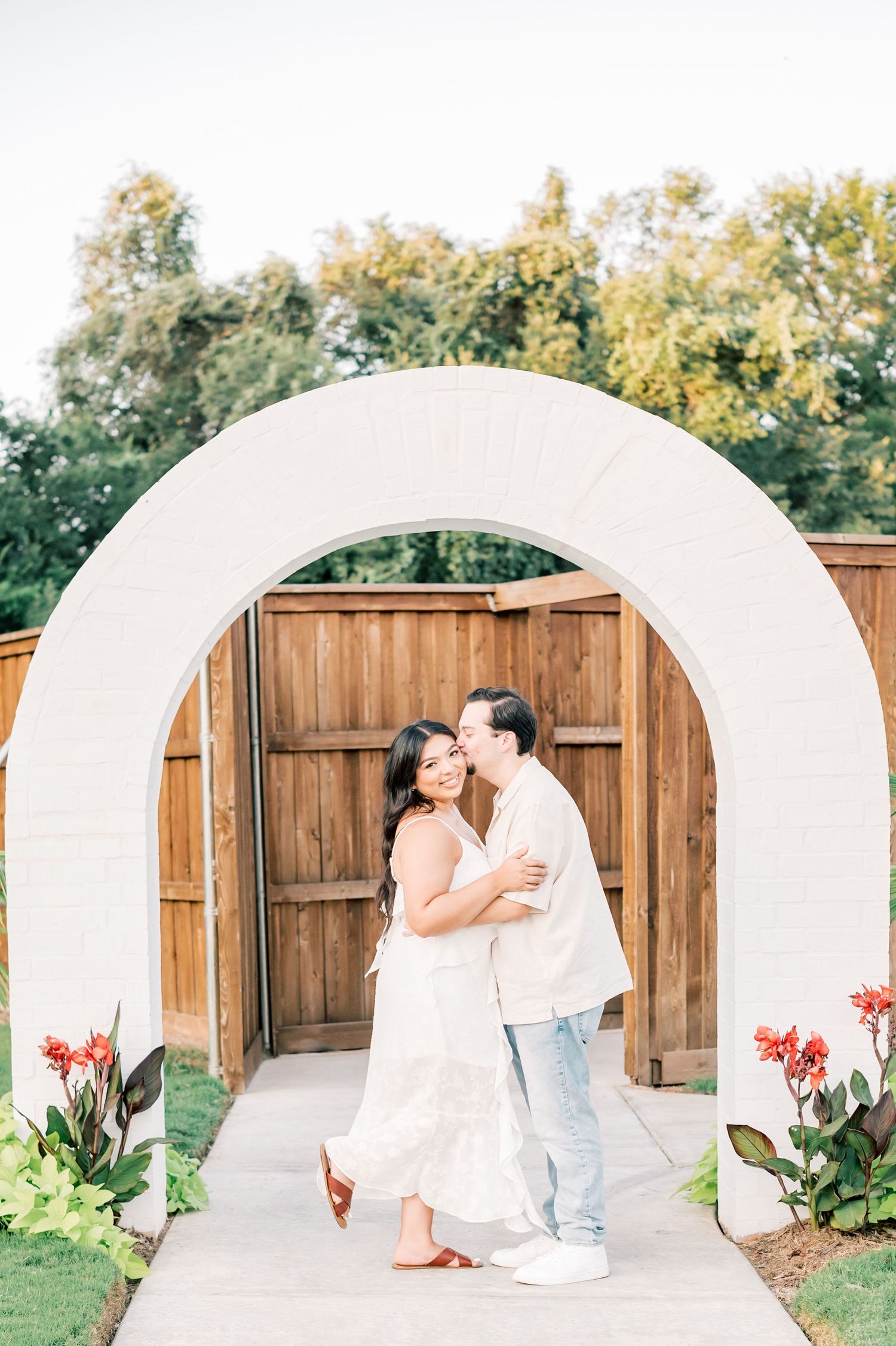 A romantic couple in white outfits shares tender moments in front of a wooden fence during an outdoor photo session.